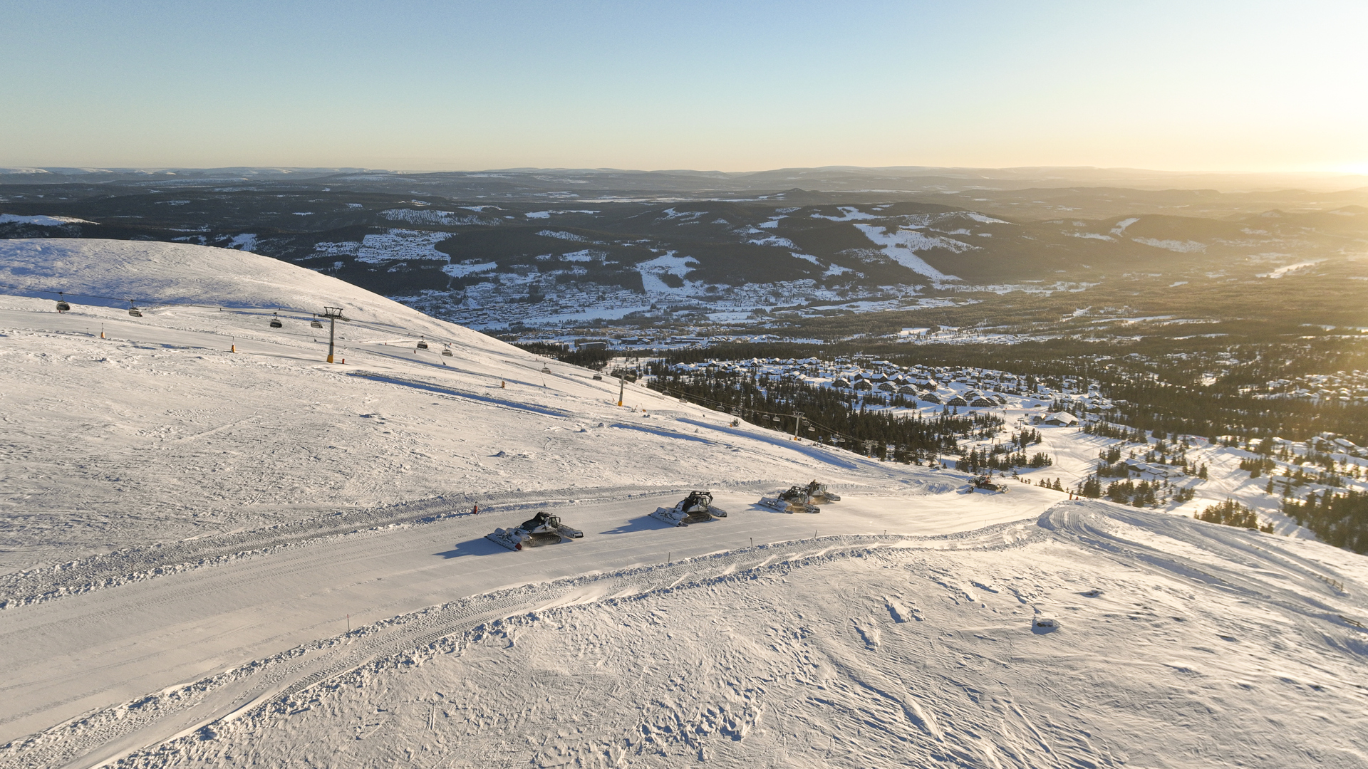 Snow-covered mountain landscape with snowmobiles on a trail, overlooking valleys and forests under a clear blue sky at sunset