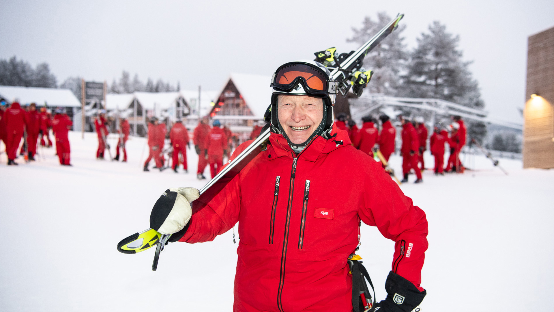 A person in a red ski jacket with skis on their shoulder is smiling at a snowy ski resort. Several people in the background are also wearing red jackets and getting ready for skiing