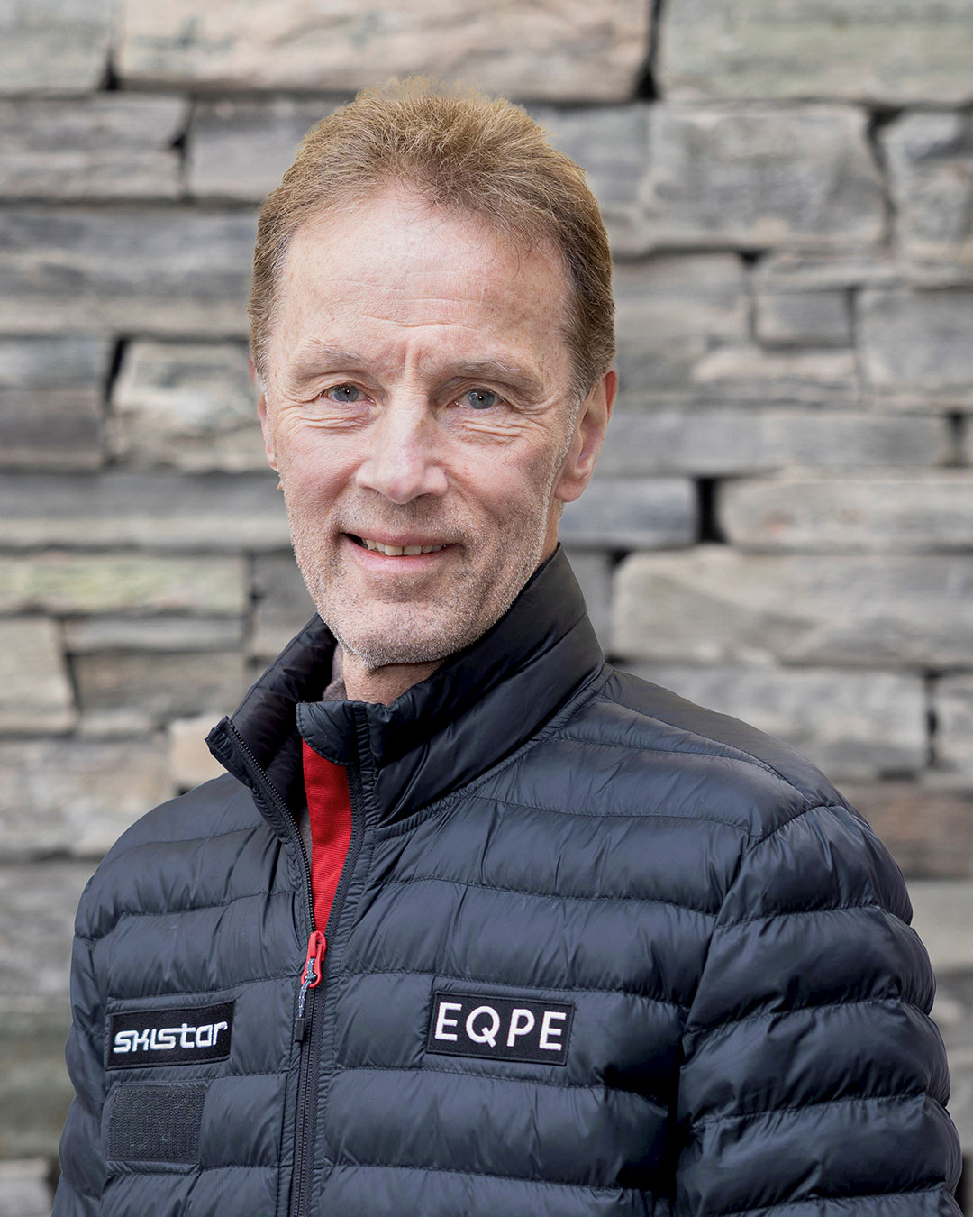 Person i svart vadderad jacka med märkena EQPE och SkiStar, står framför en stenmur