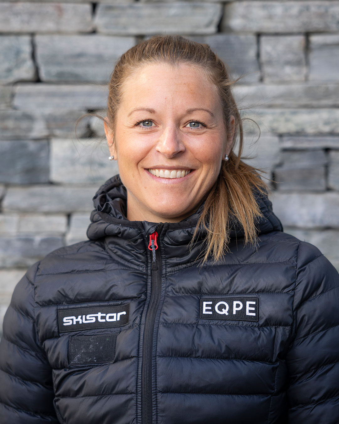 Kvinna i svart dunjacka med loggor från SkiStar och EQPE, stående framför en stenmur. Hon ler och har blont hår