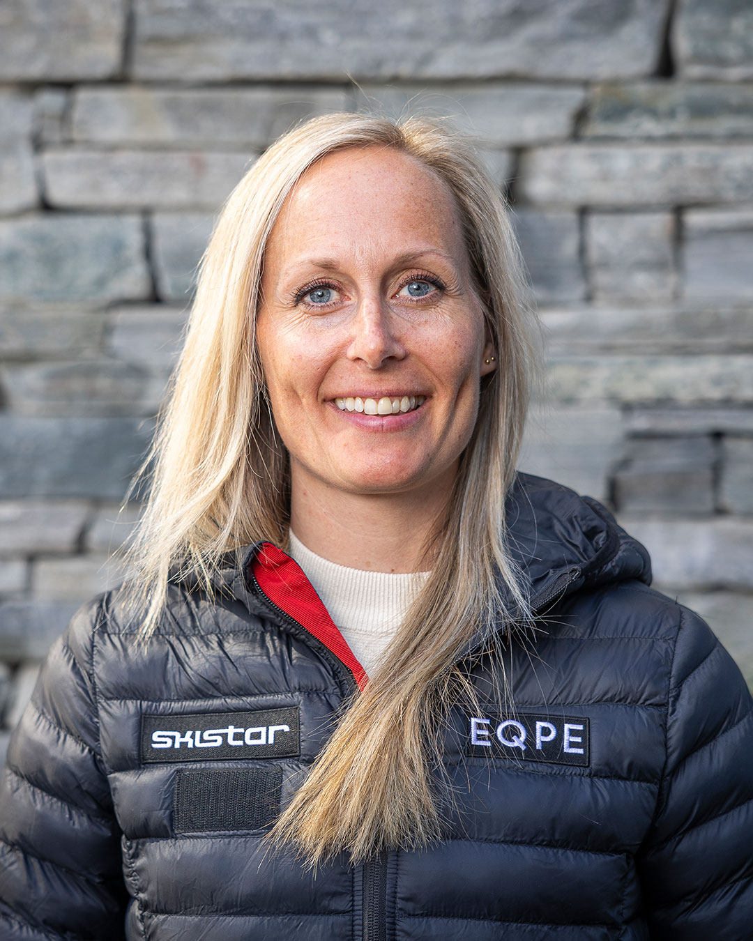 Kvinna med långt blont hår klädd i en svart dunjacka med logotyperna SkiStar och EQPE, står framför en stenmur