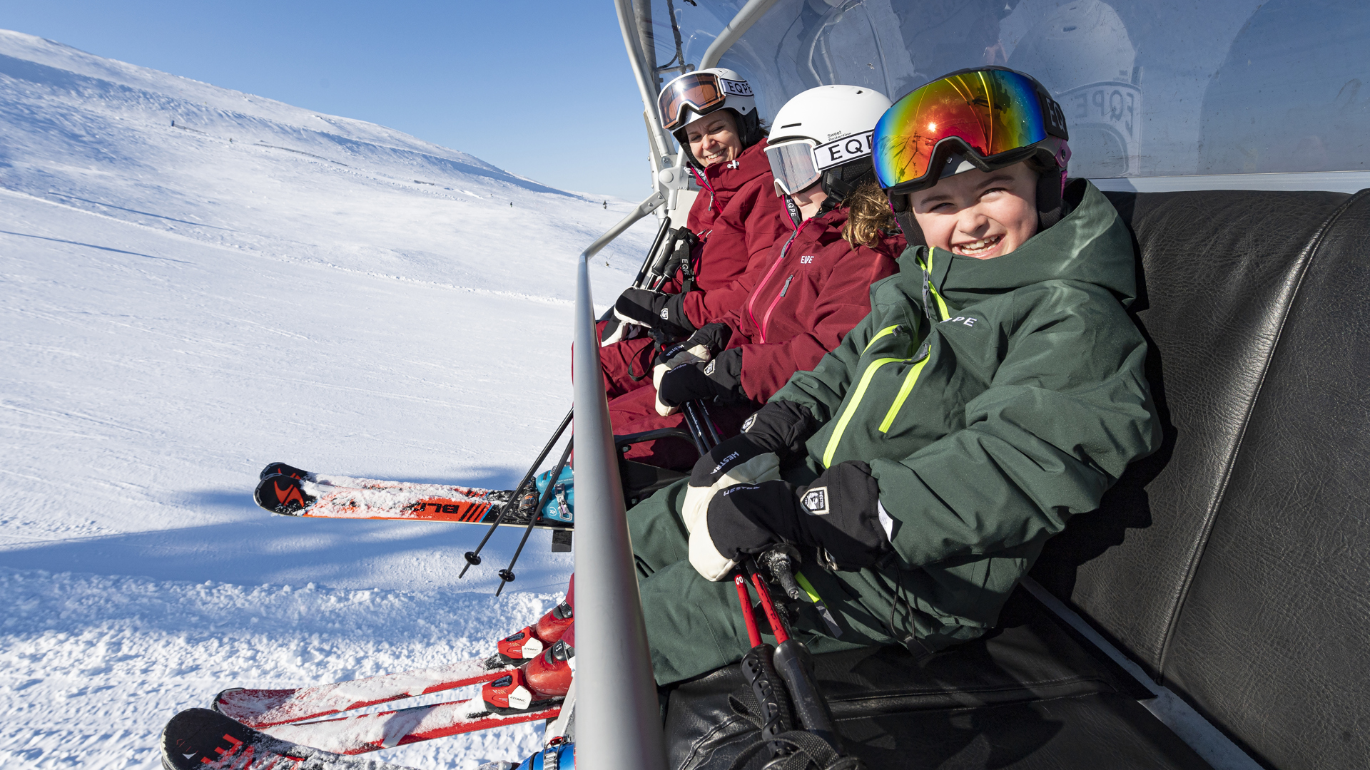 Fyra personer i skidkläder sitter i en stollift på en solig dag i ett snötäckt fjällandskap