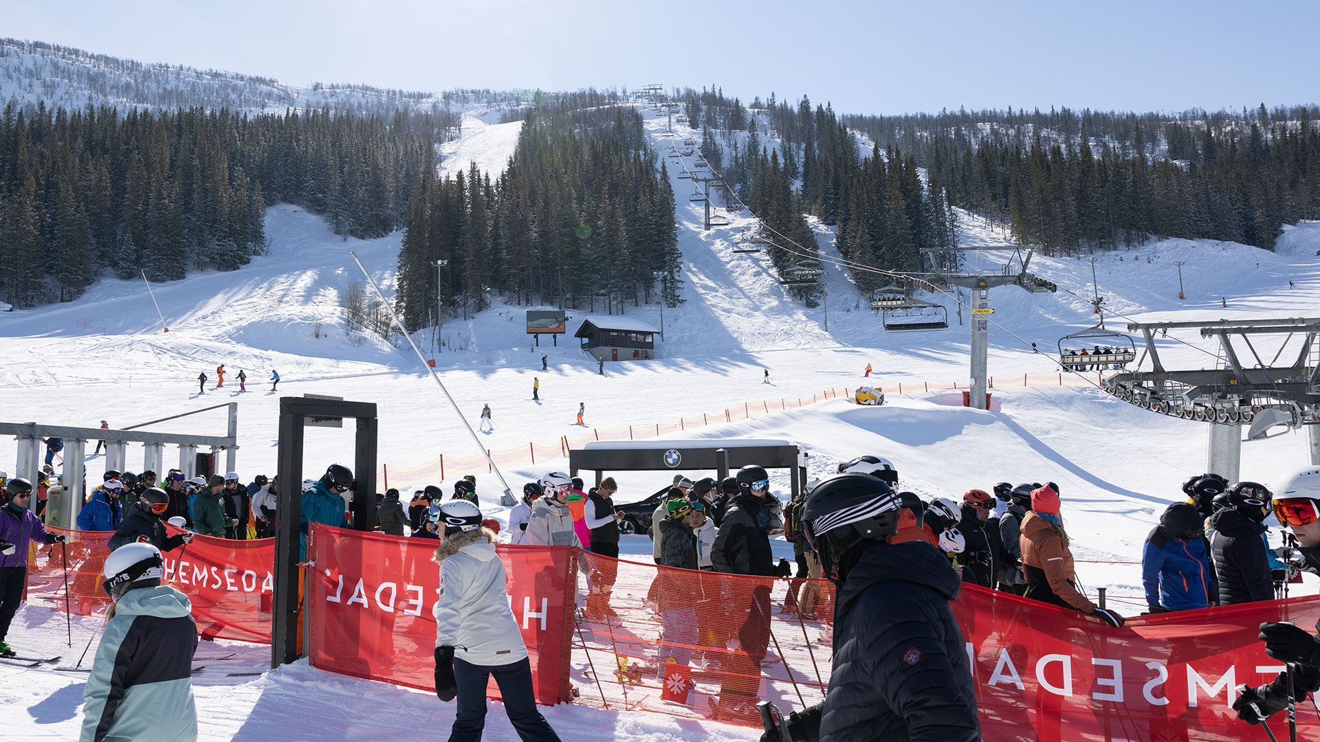 Skiers in Hemsedal, Norway, wait by a lift on a sunny winter day with snow-covered mountains and pine trees in the background