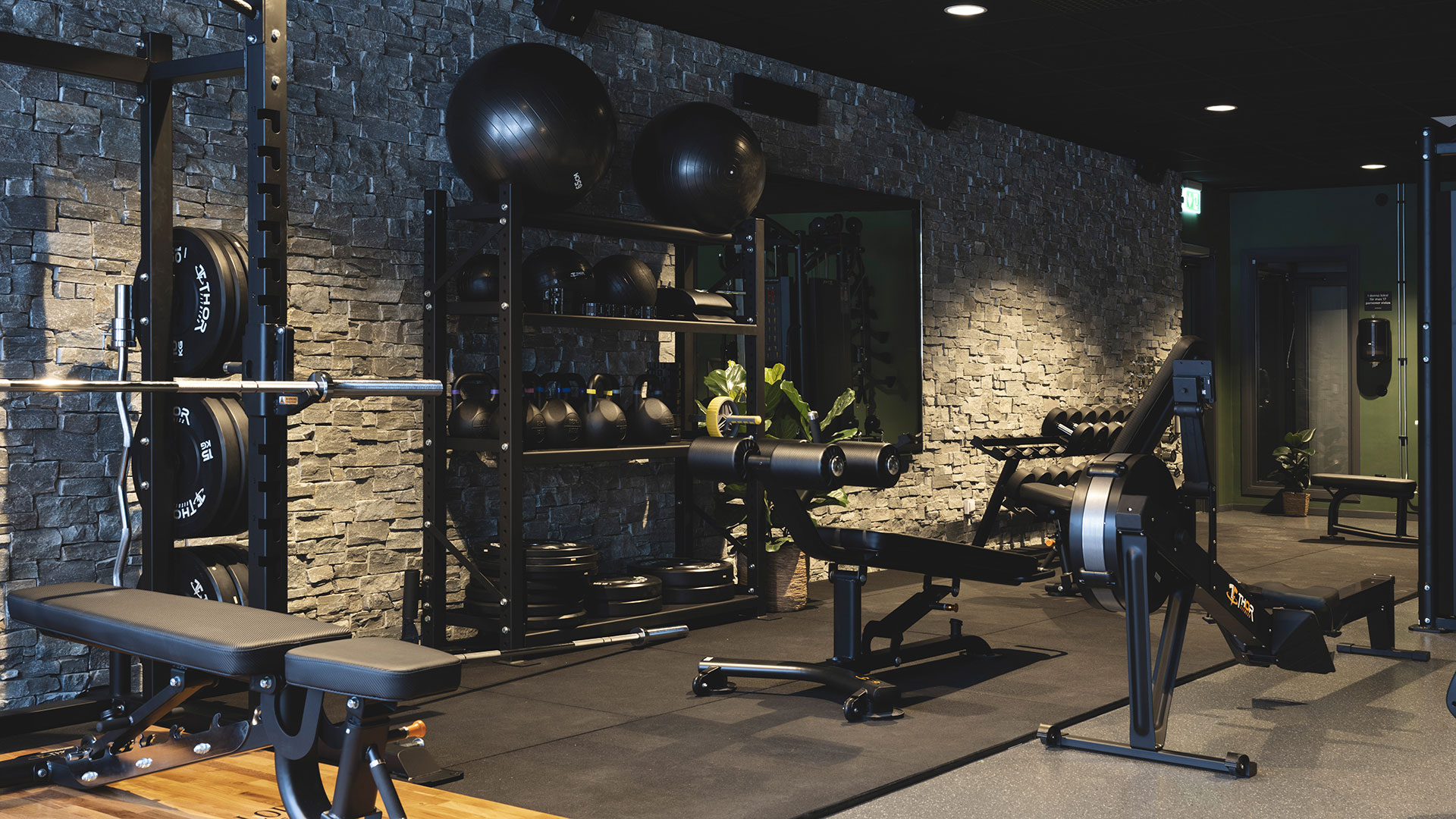 Modernt gym med styrketräningsutrustning, inklusive bänkar, hantlar och skivstänger, mot en vägg av sten
