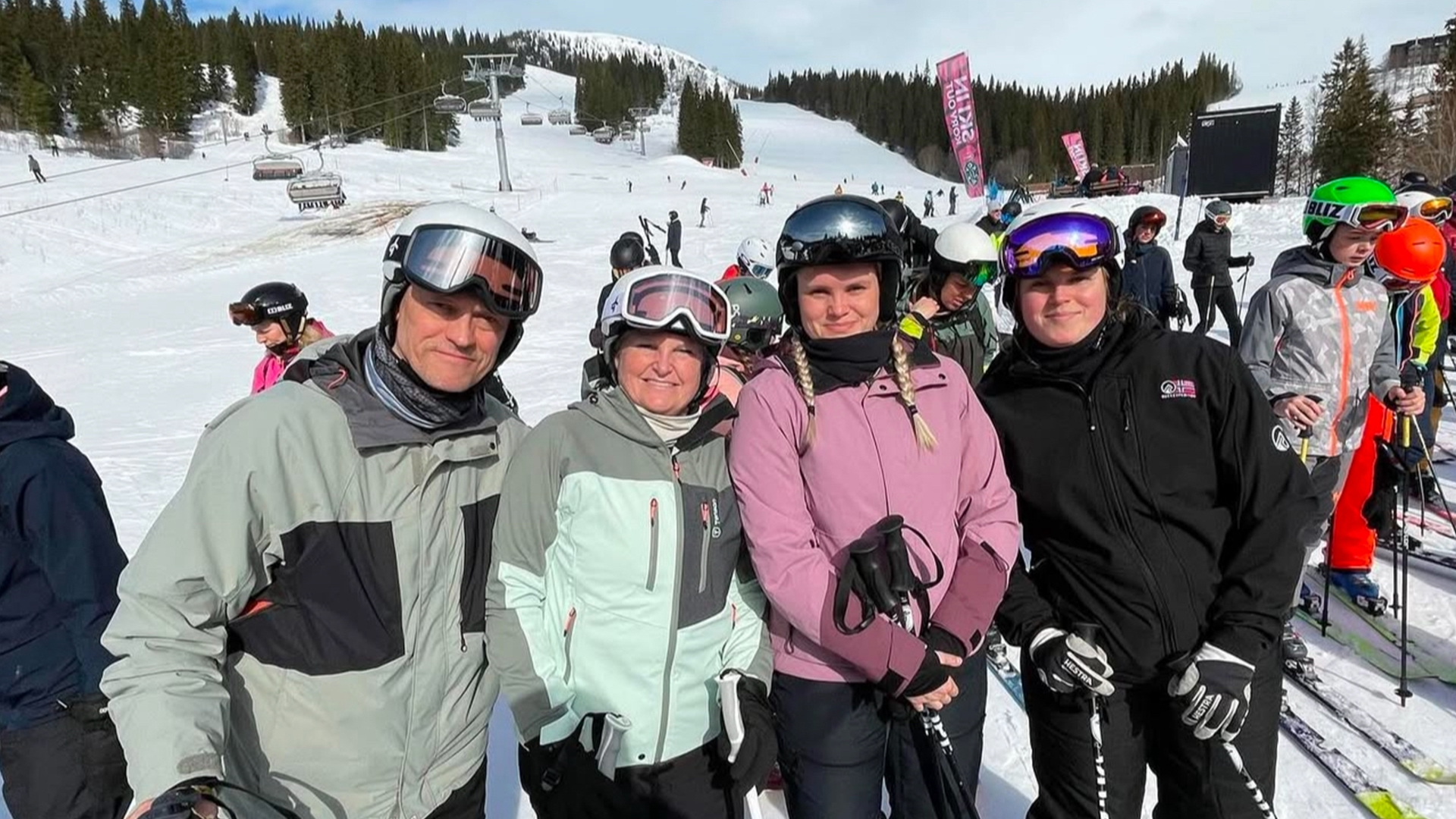 Fyra personer på skidsemester i en snöig alpin miljö med skidlift och andra skidåkare i bakgrunden