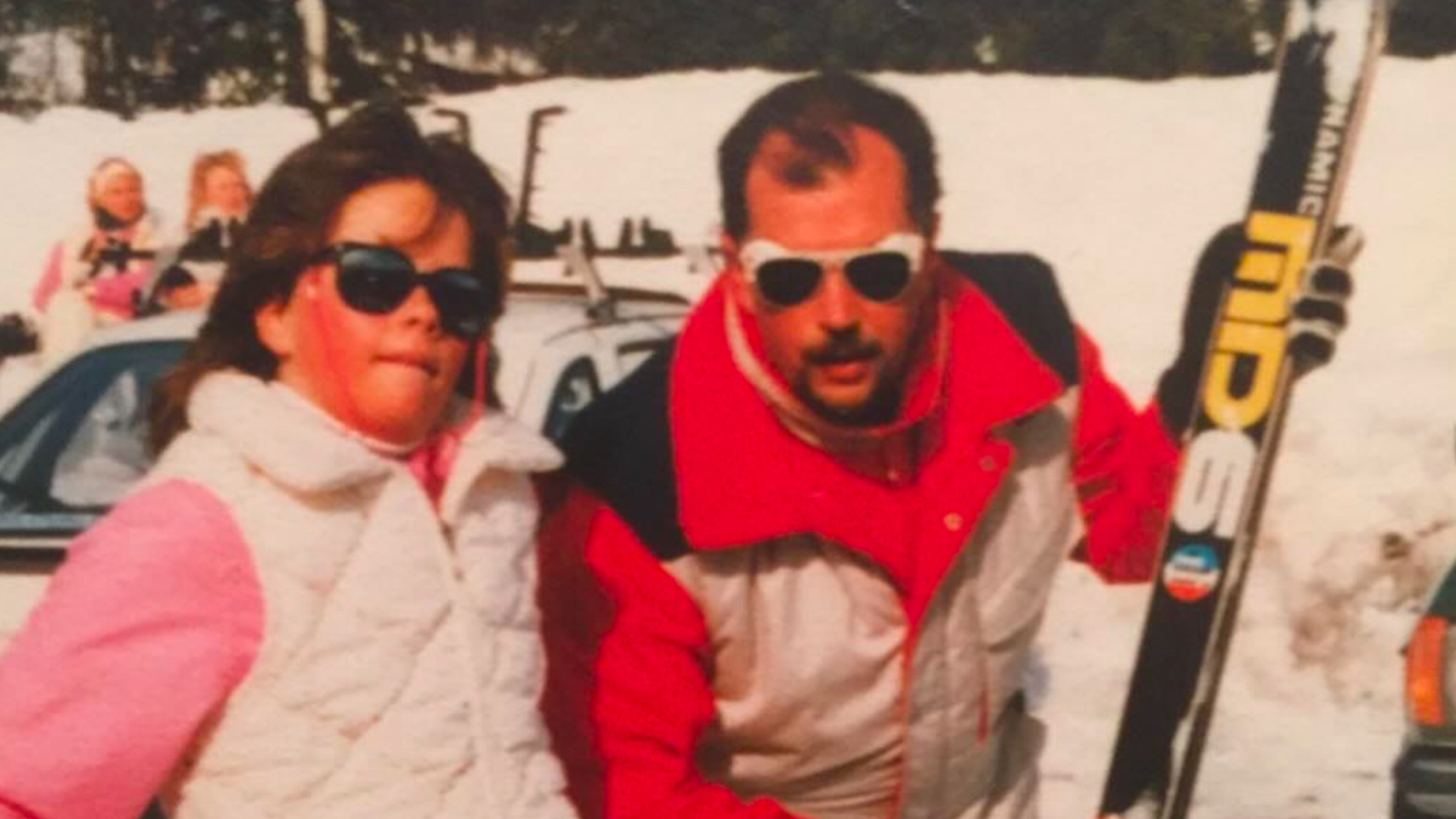 Man och barn i skidkläder med solglasögon, står framför en bil med skidor, på en snötäckt plats