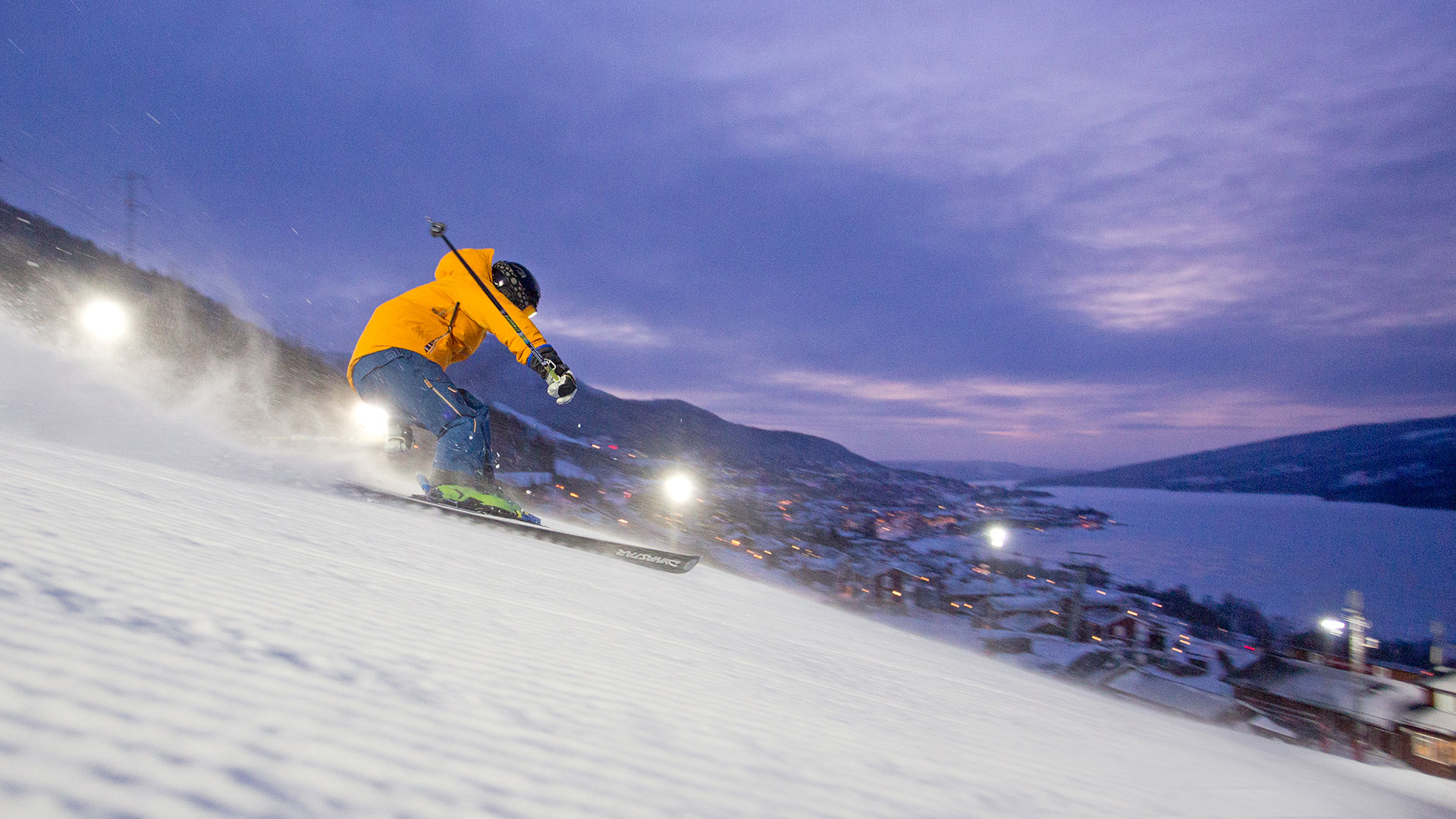 Person åker skidor nerför en snöig backe i Åre, Sverige, vid skymning med staden och sjön i bakgrunden