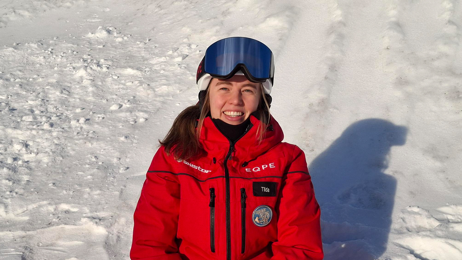 Person i röd skidjacka och skidglasögon sitter i snön och ler, med vintrigt landskap i bakgrunden