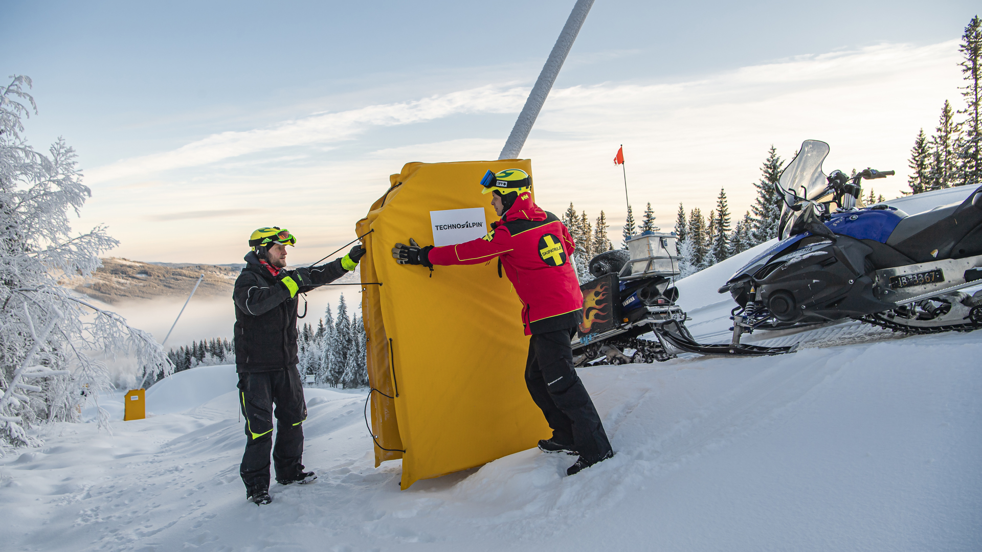 Två personer på en snöig fjällsluttning installerar en gul skidbacksskyddsmatta nära en skoter i en vintrig landskap