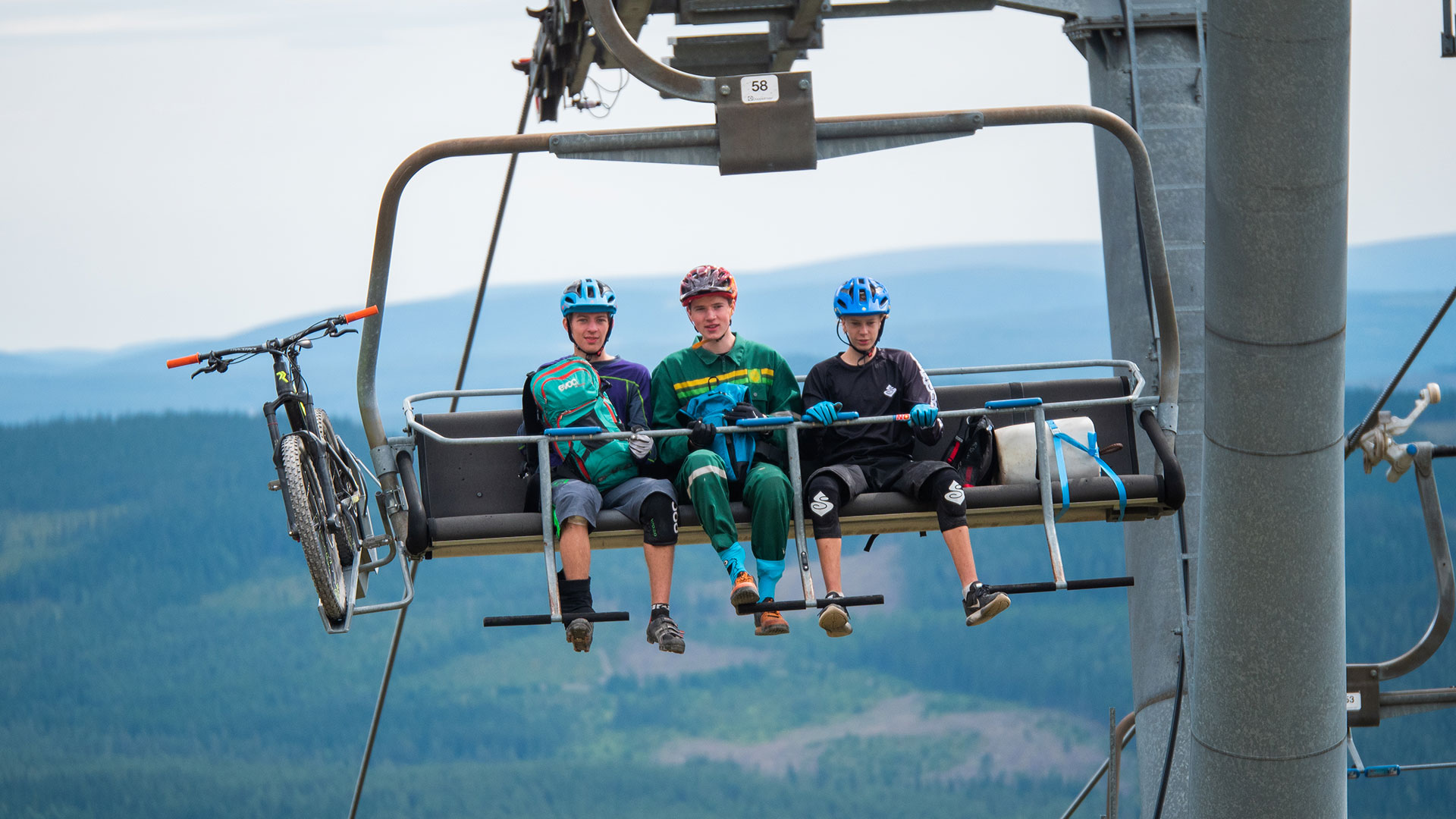Tre personer med cykelhjälmar åker en stollift uppför ett berg med cyklarna på sidan, med en skog och berg i bakgrunden