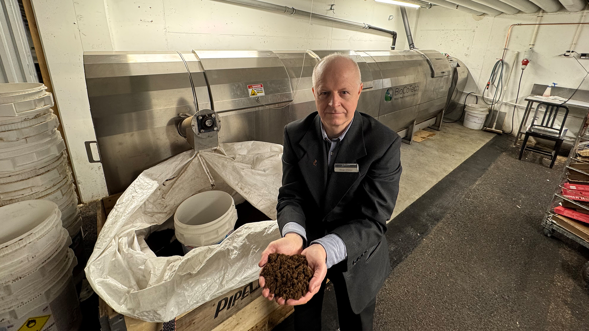 Man i fabriksmiljö håller kompostmaterial framför stor maskin med etiketten "BioCoTech", omgiven av hinkar och pallar