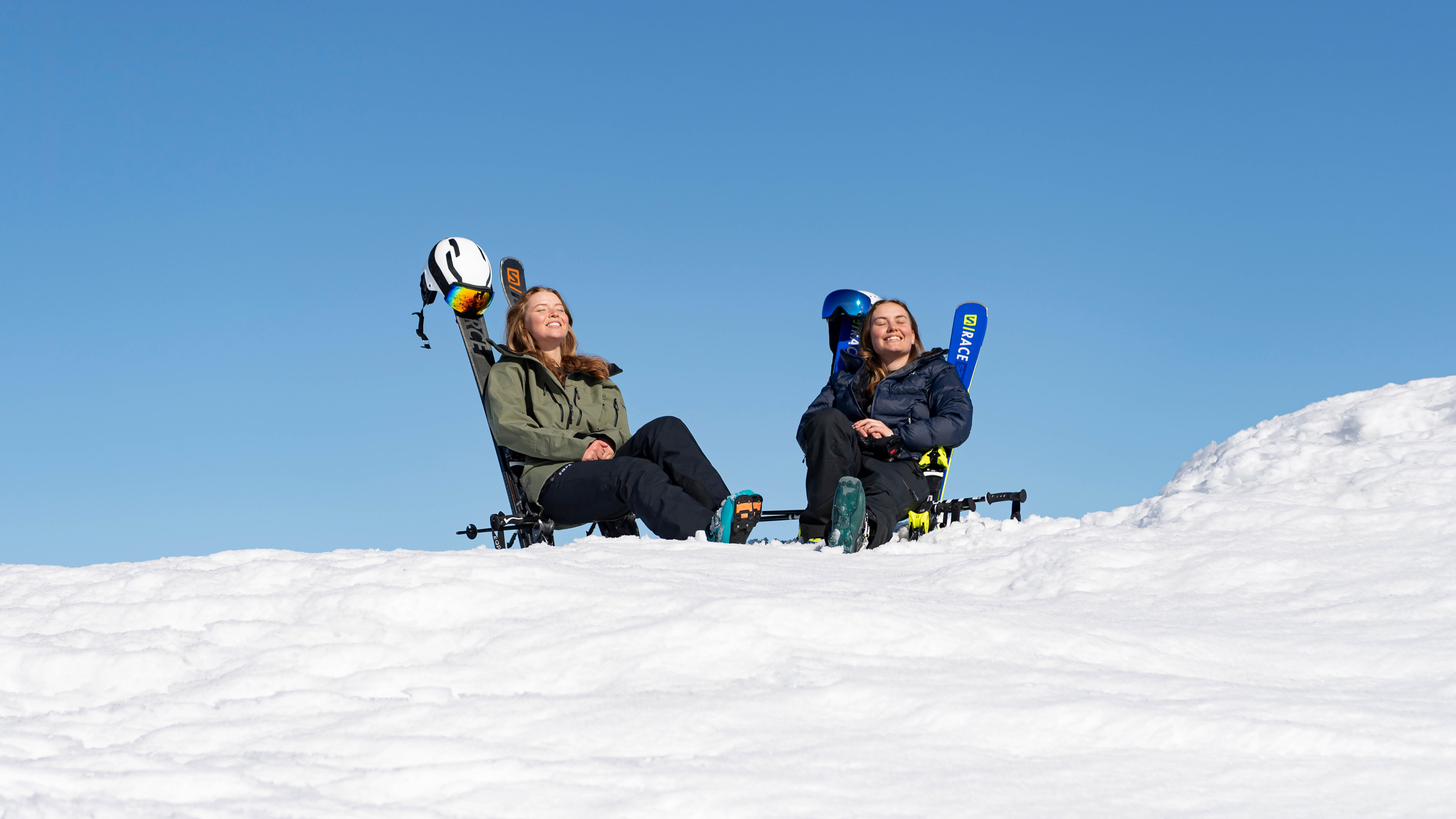 Två personer sitter i solstolar i snön under en klarblå himmel. De har skidutrustning, inklusive hjälmar och skidor, bredvid sig och ser glada och avslappnade ut