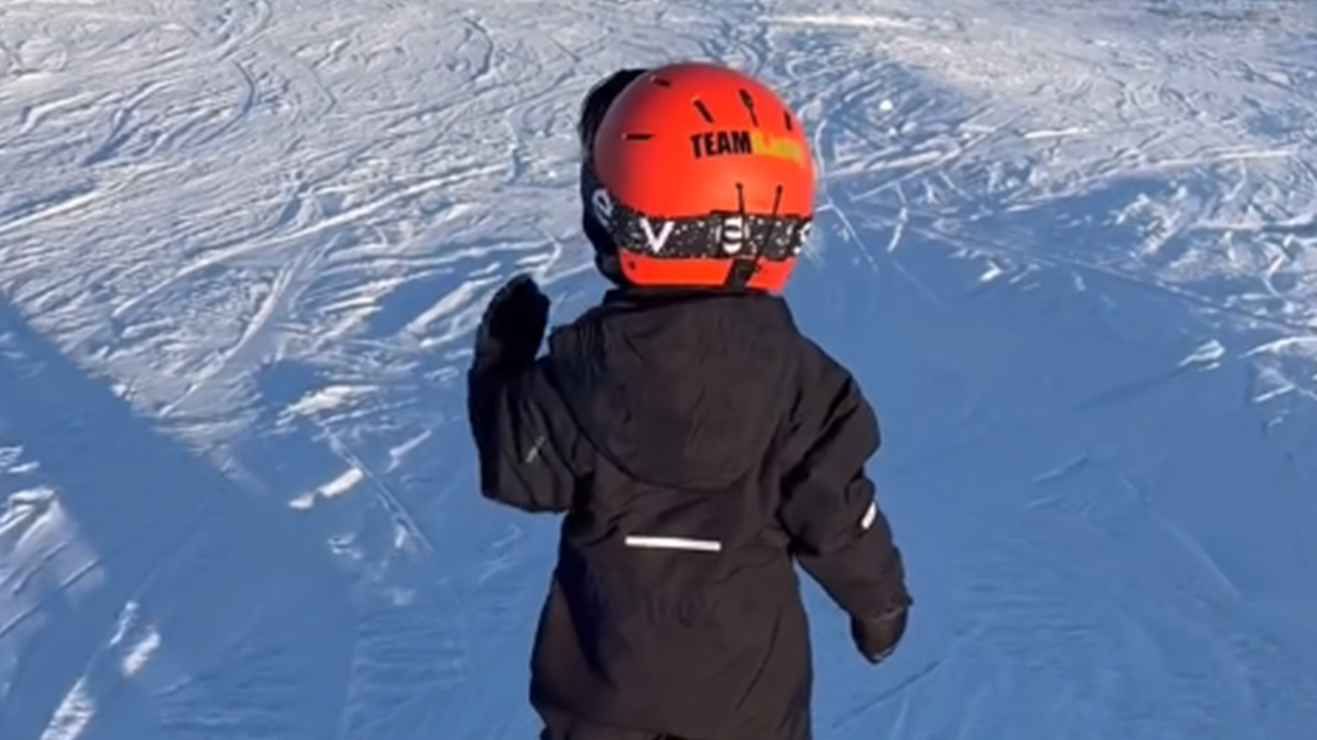 Barn åker skidor på snötäckt backe, bärande röd hjälm och svart overall