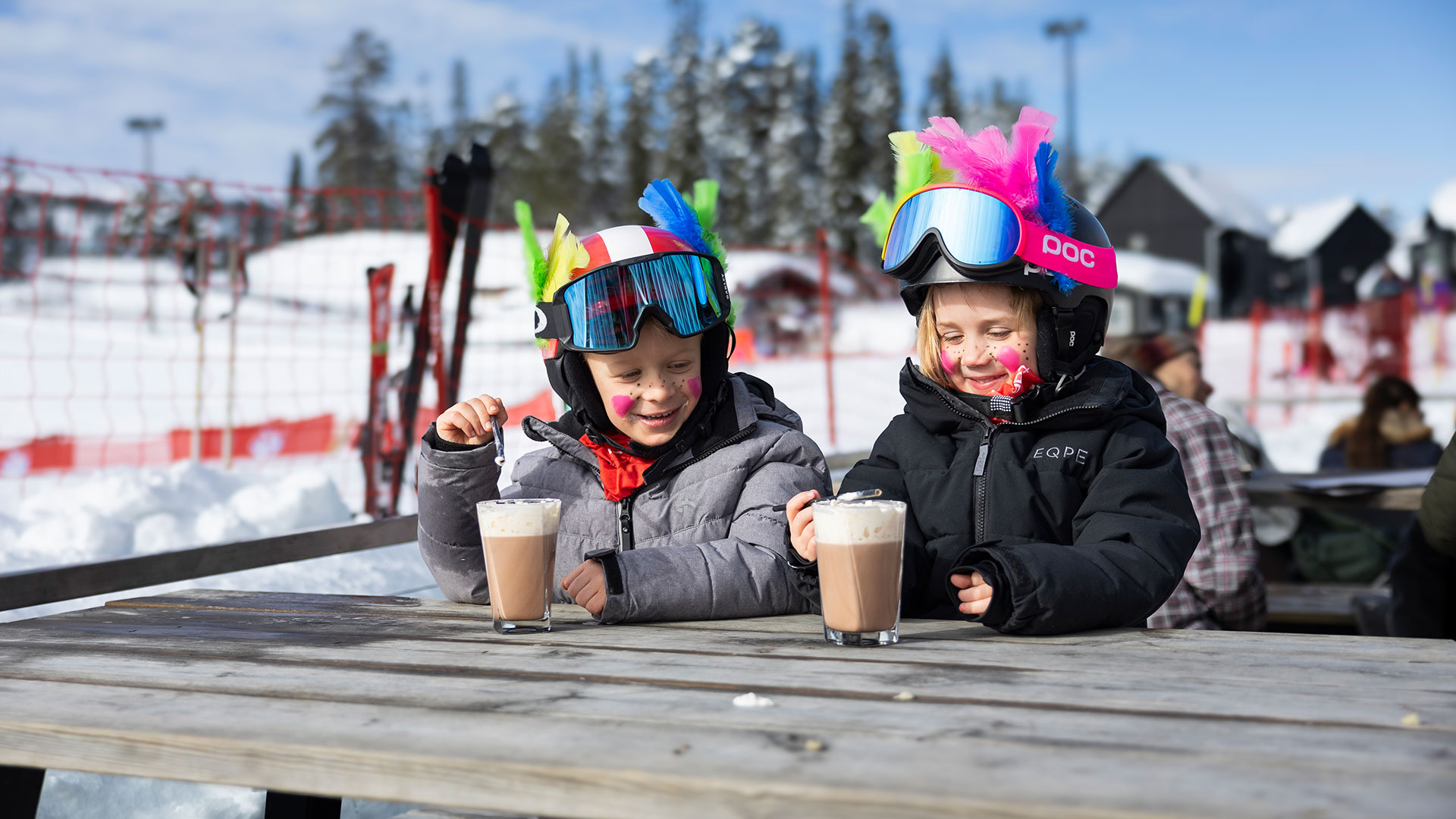 Två barn i skidutrustning dricker varm choklad vid ett utomhusbord i en skidort. Snö och skidbackar syns i bakgrunden