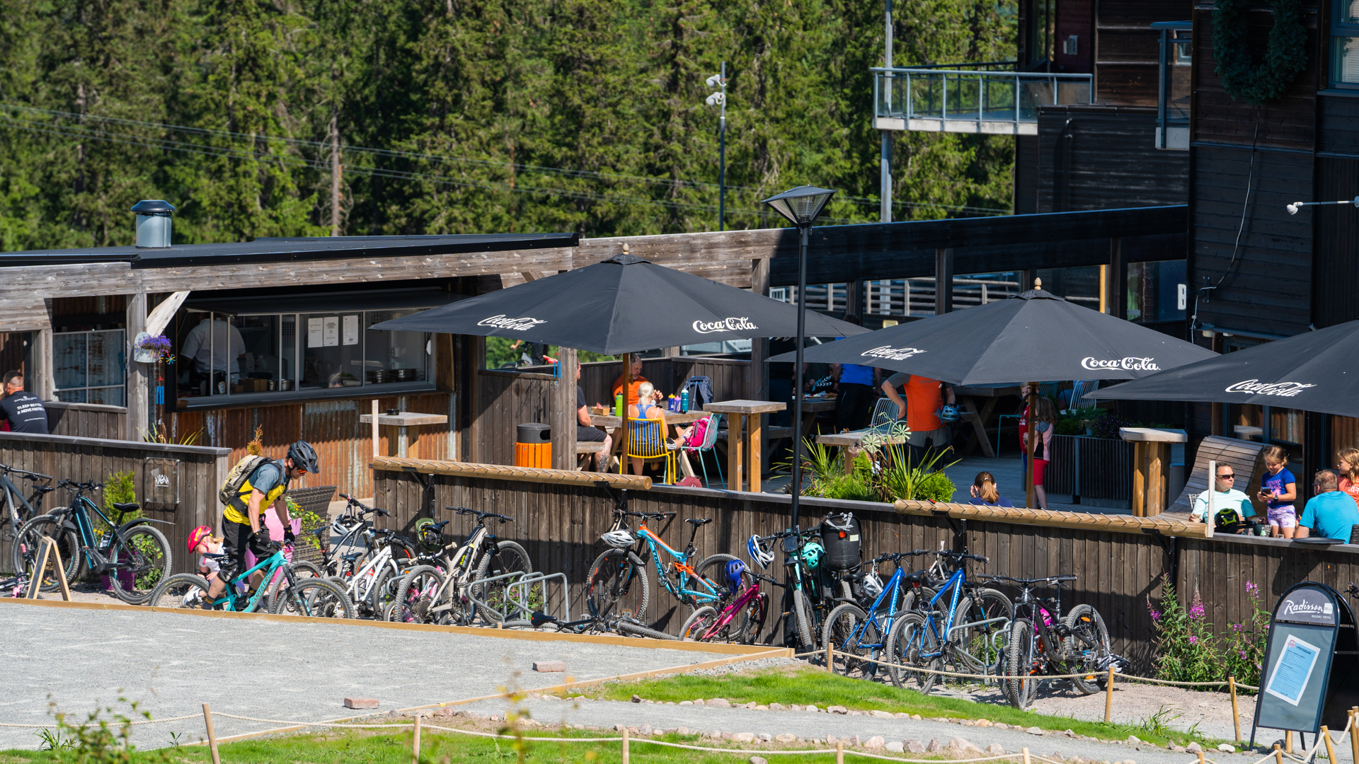 Cyklar parkerade utanför ett café i en skogsmiljö med trätak och parasoller, människor sitter och fikar