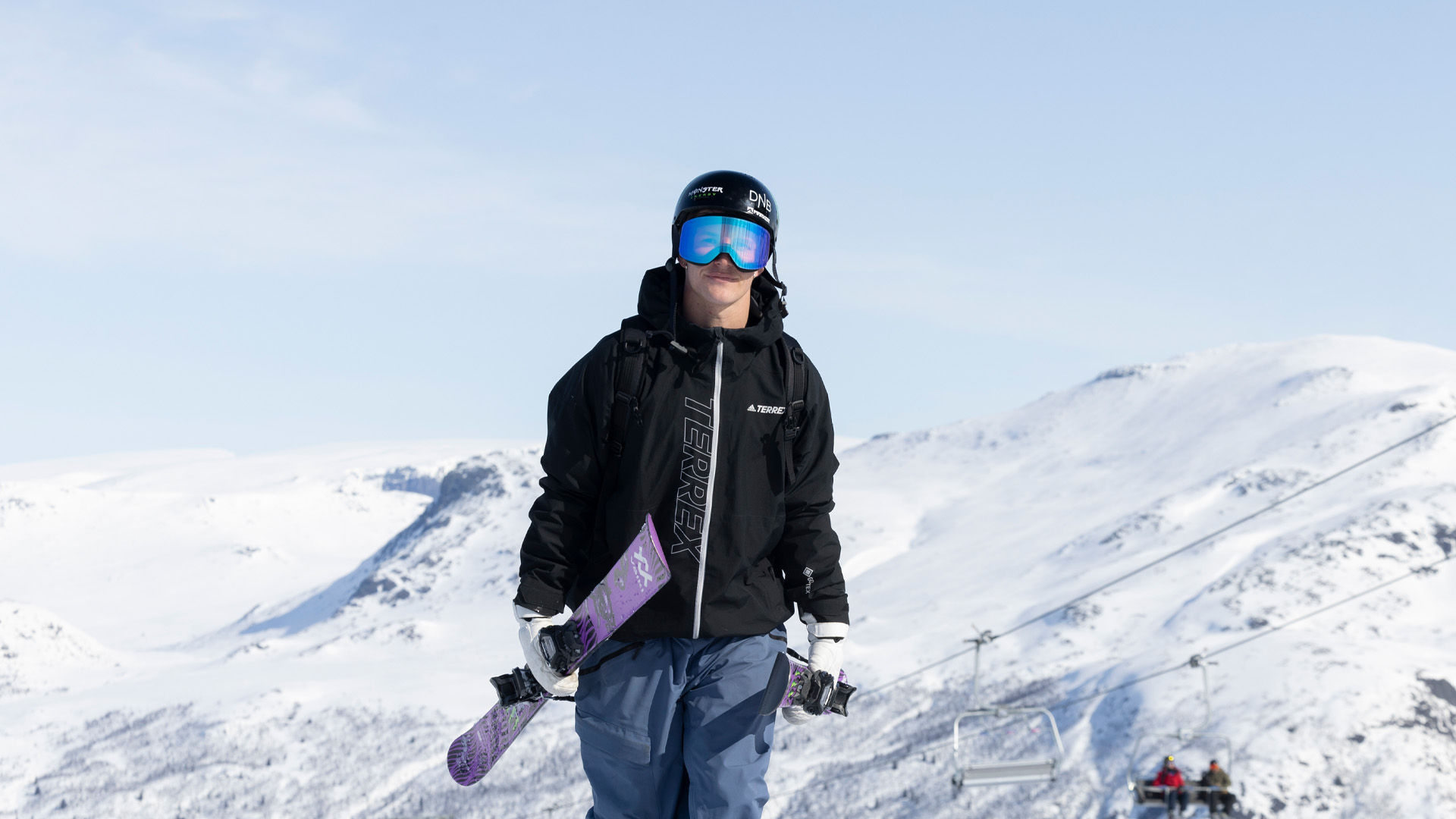 Person klädd för skidåkning med hjälm och glasögon håller en snowboard mot en bakgrund av snötäckta berg. En skidlift syns i bakgrunden
