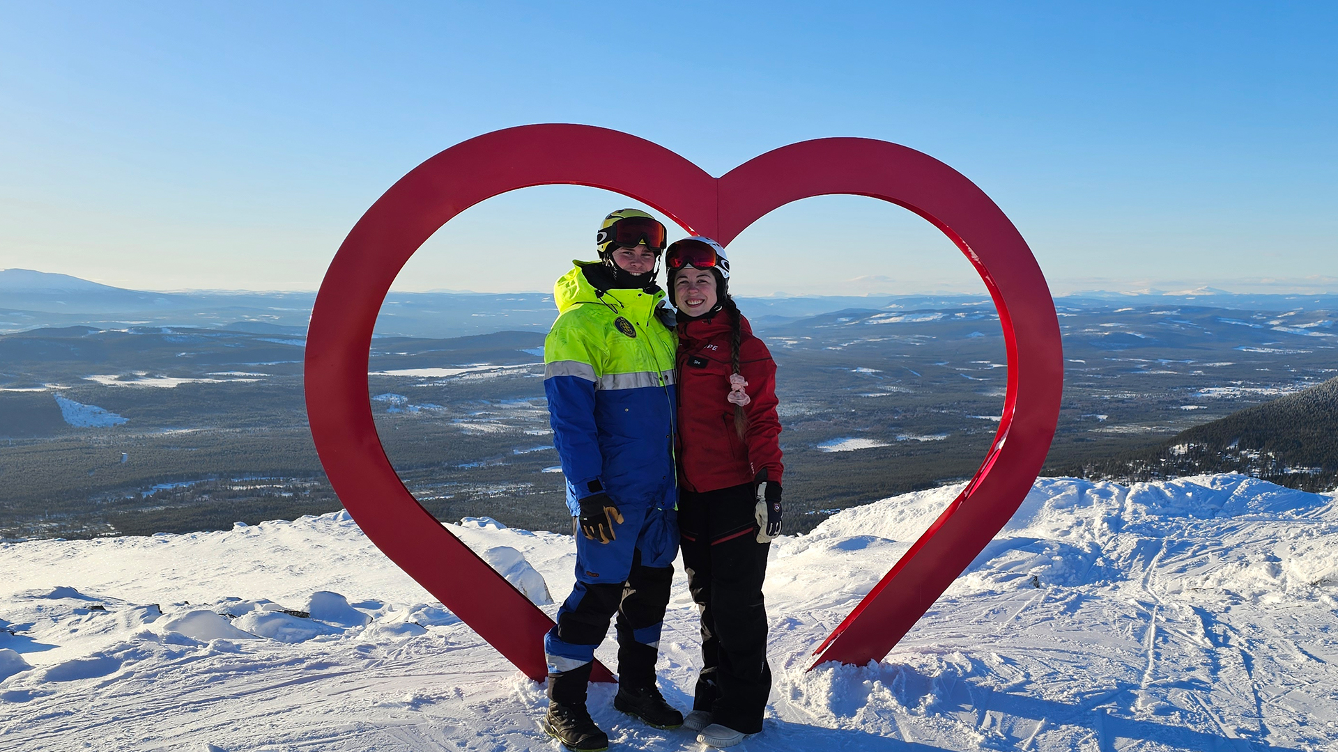 People are posing by a heart-shaped artwork on a snowy mountaintop with a vast view in the background, wearing ski-inspired clothing