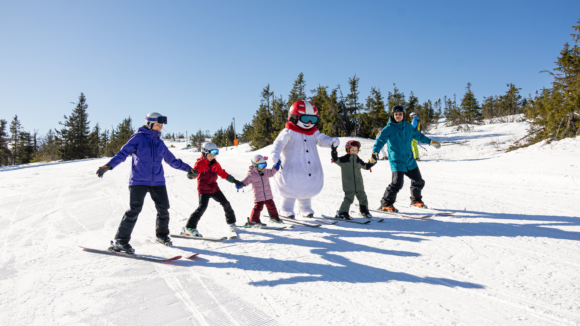 Familj åker skidor med en snögubbe-maskot i fjällen, en solig vinterdag med snöklädda träd i bakgrunden