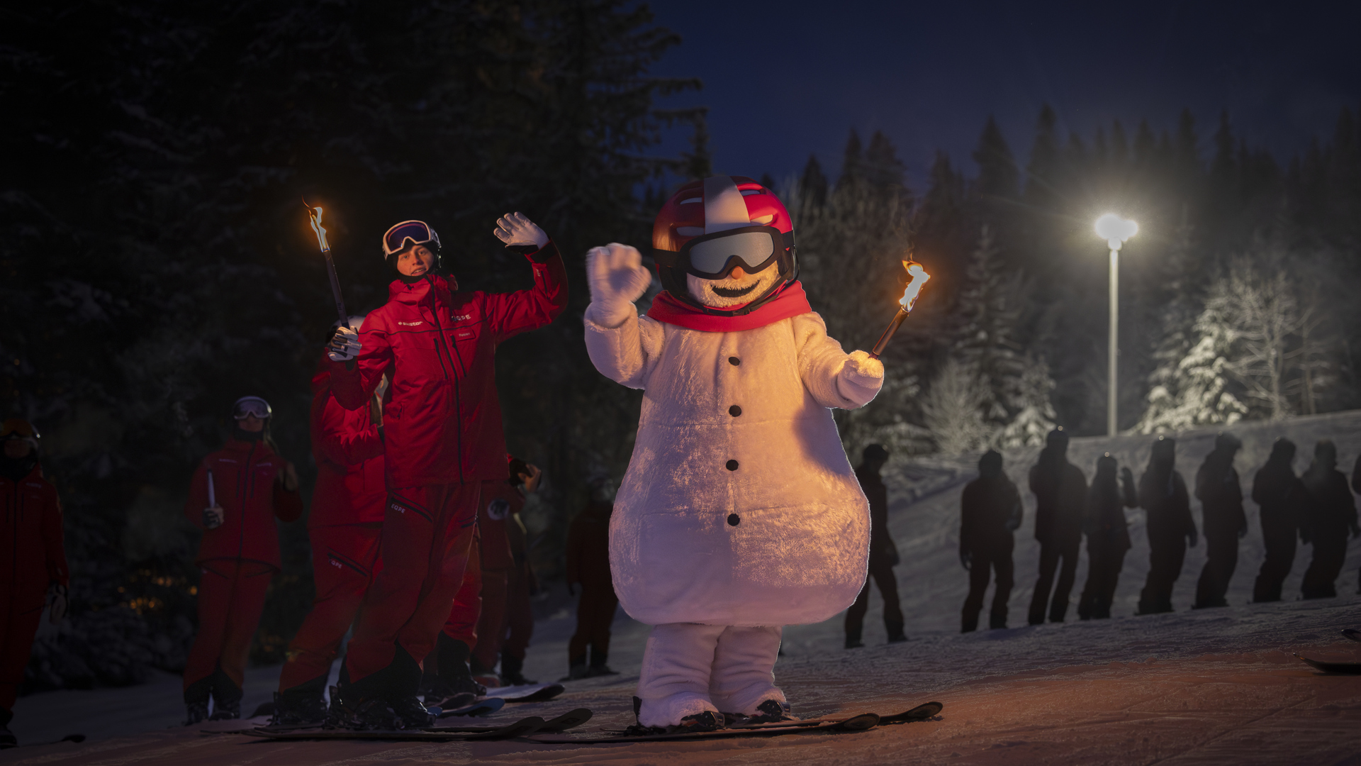 Snögubbe med skidutrustning håller facklor omgiven av skidlärare i röda dräkter under en kvällsshow på snötäckt skidbacke
