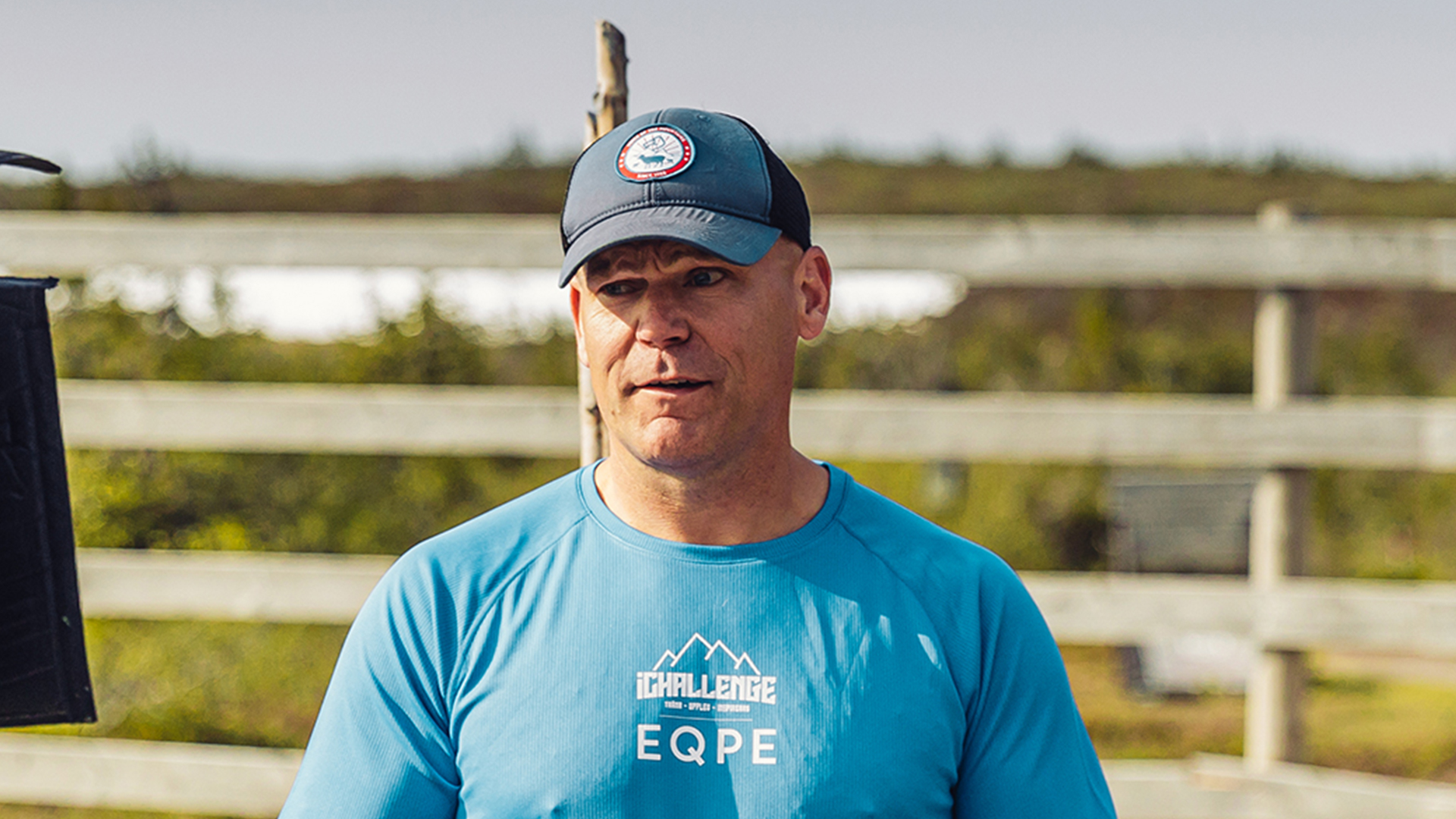 Man i blå t-shirt och keps utomhus, vid stängsel och grönområde