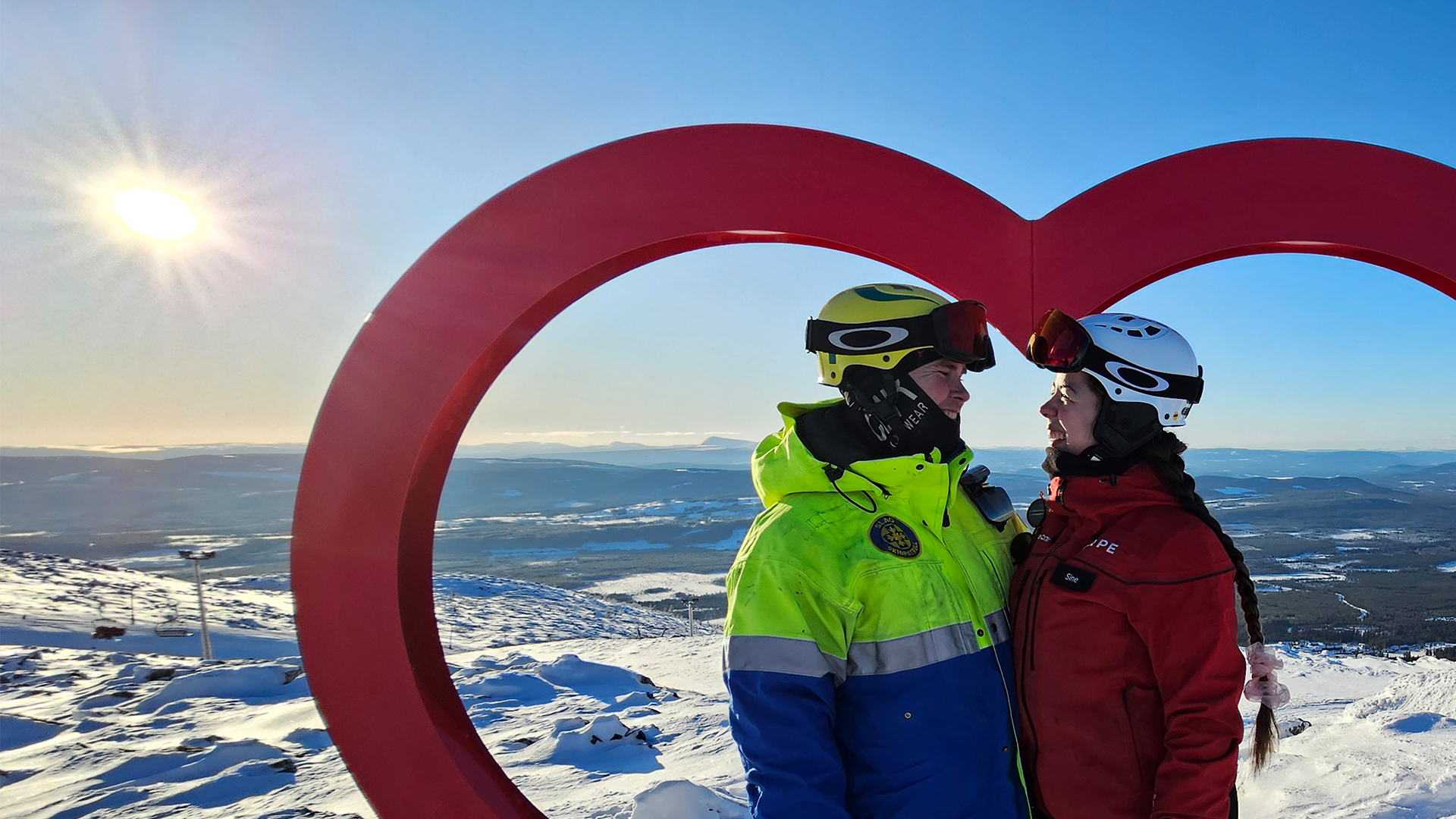 Två personer i skidkläder står under en hjärtformad installation på en snötäckt bergstopp med utsikt över ett solbelyst vinterlandskap i Sverige