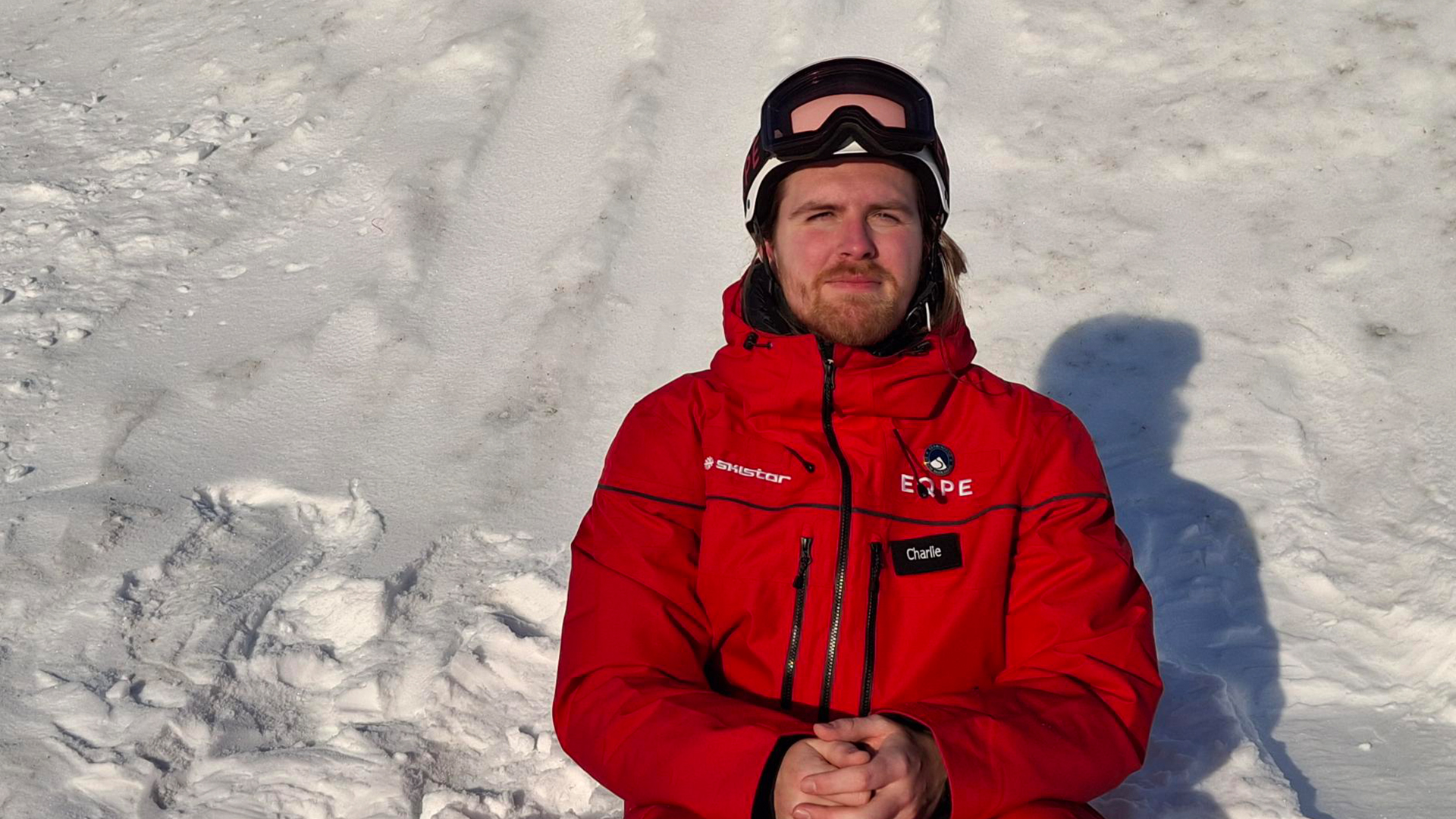 Person i röd jacka sitter på snö, utrustad med skidglasögon och hjälm, mot vinterlandskap