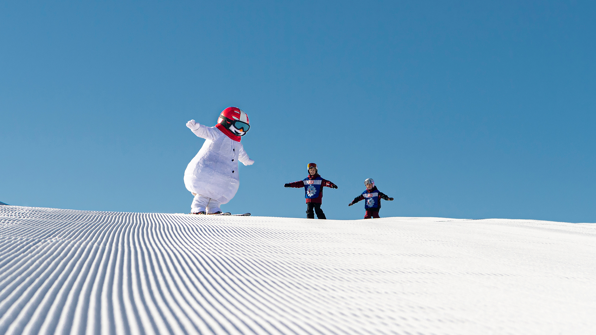 Snögubbe med röd hjälm åker skidor med två barn på en snötäckt pist under klarblå himmel