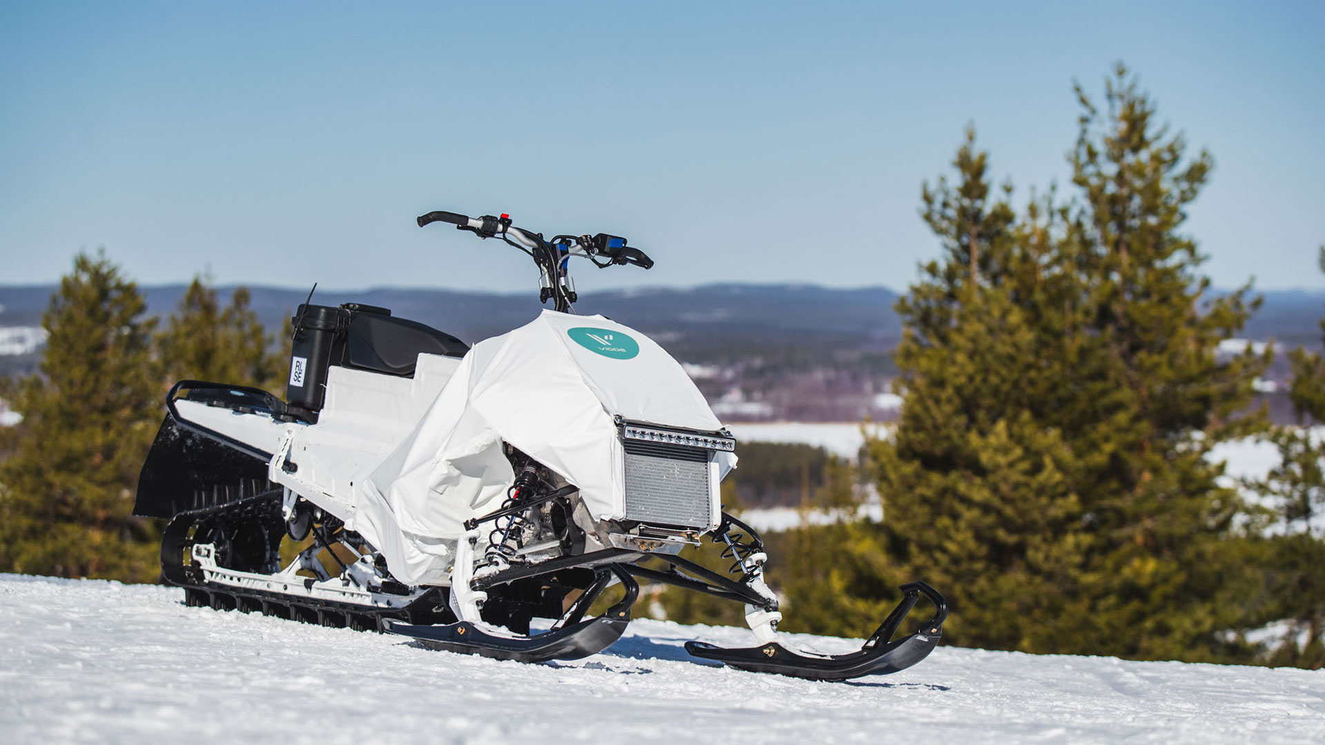 Vit snöskoter parkerad i vinterlandskap med snötäckta marker och skog i bakgrunden, under klar himmel