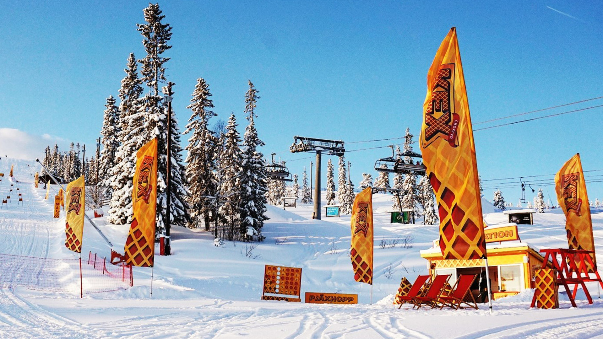 Snöigt landskap vid skidort med färgglada flaggor, stolliftar i bakgrunden och klarblå himmel i Åre, Sverige