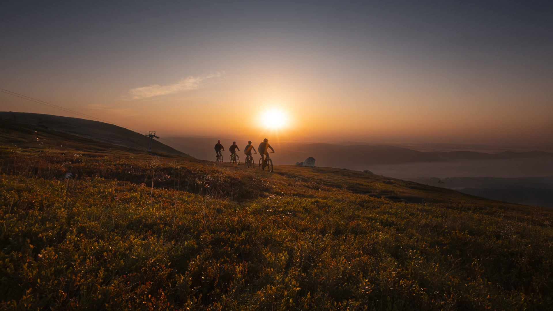 Radfahrergruppe bei Sonnenuntergang auf einem Berggipfel, mit dramatischem Himmel und weitläufiger Landschaft im Hintergrund