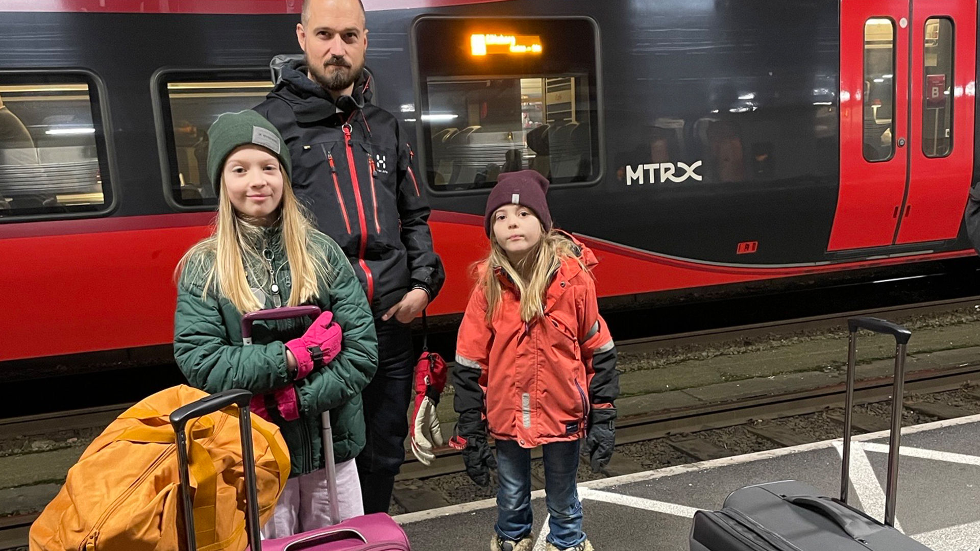 Familj väntar på ett MTRX-tåg på en järnvägsstation med resväskor