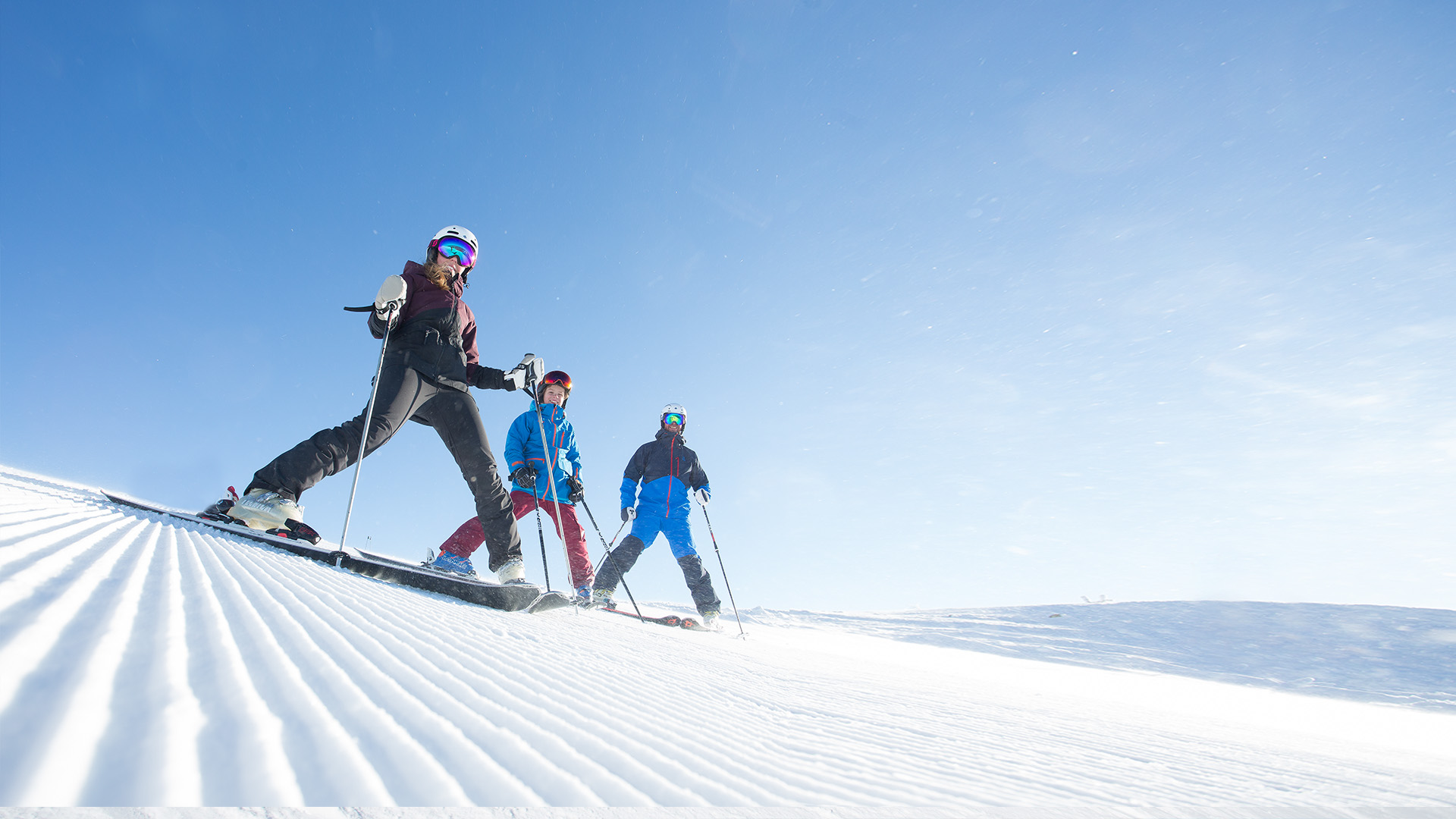 Tre personer står på ski ned ad en pist i solskinsvejr med en klarblå himmel