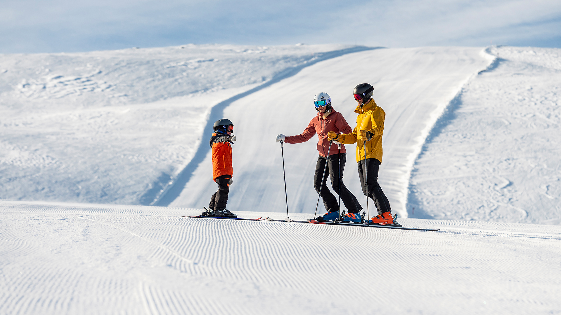 Tre personer åker skidor på en solig skidbacke med snöklädda berg i bakgrunden. De bär färgglada skidkläder och hjälmar
