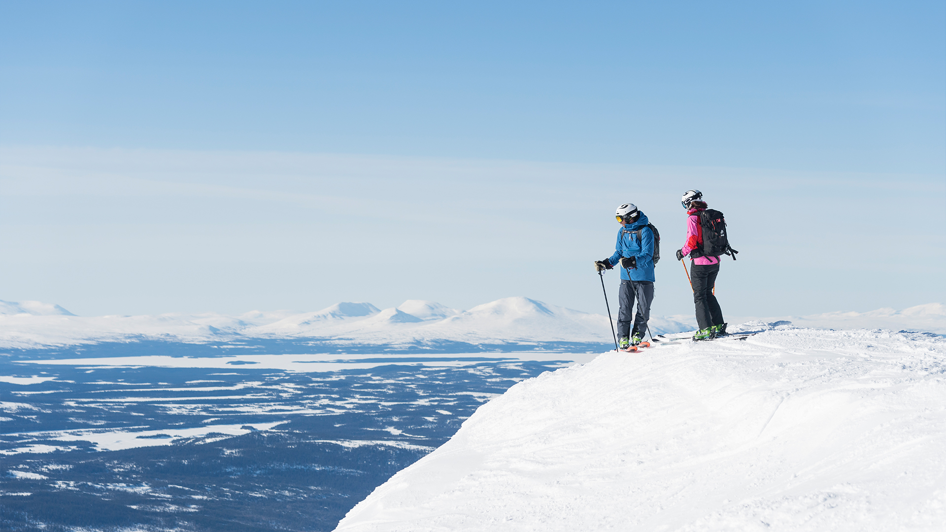 Två skidåkare på toppen av ett snötäckt fjäll i Sverige, med vidsträckt utsikt över berg och dalar under en klarblå himmel