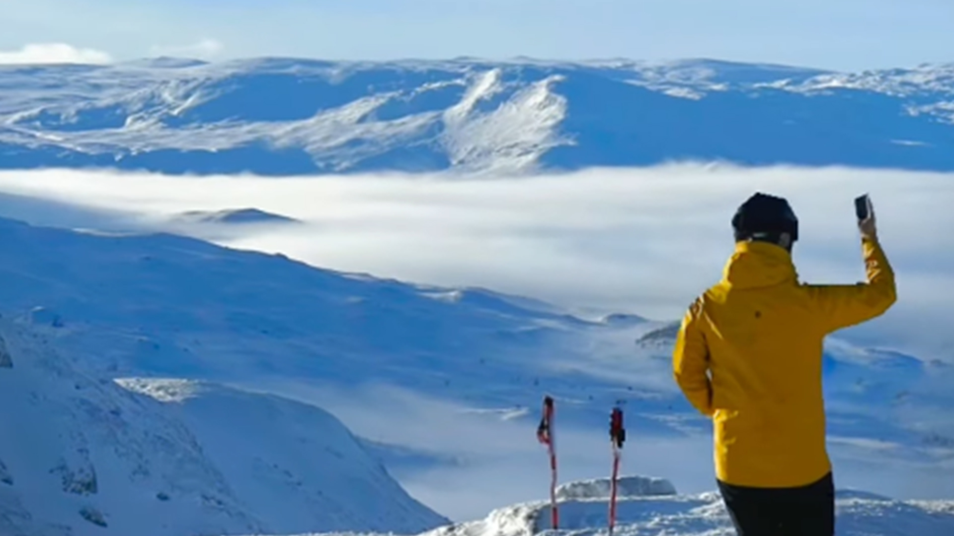 Person i gul jacka tar foto på snötäckt fjällandskap med skidor i bakgrunden, vintersport i Sverige