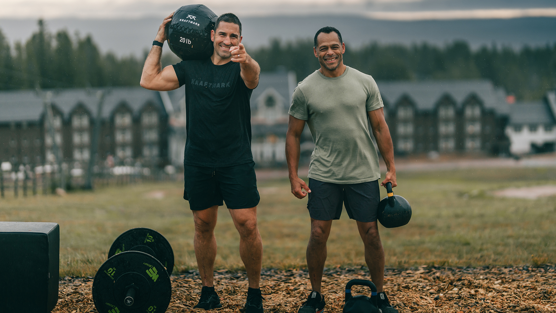 Två män tränar utomhus med vikter på en grusplan, med skog och byggnader i bakgrunden. Den ena mannen håller en medicinboll och pekar mot kameran, den andra har en kettlebell i handen