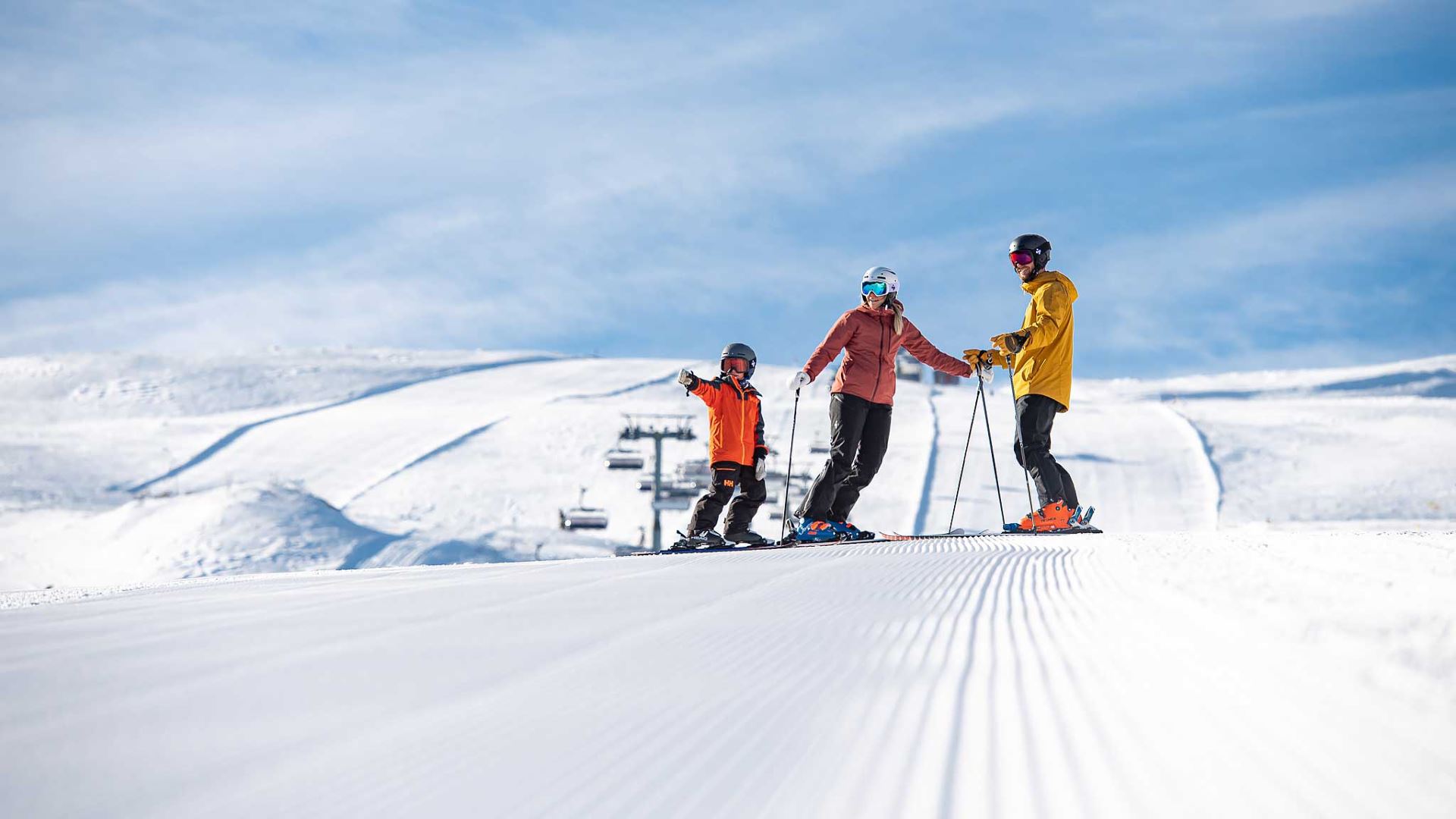 Tre skiløbere klædt i farvestrålende jakker kører ned ad en pist i et snedækket bjerglandskab med blå himmel og skilifte i baggrunden