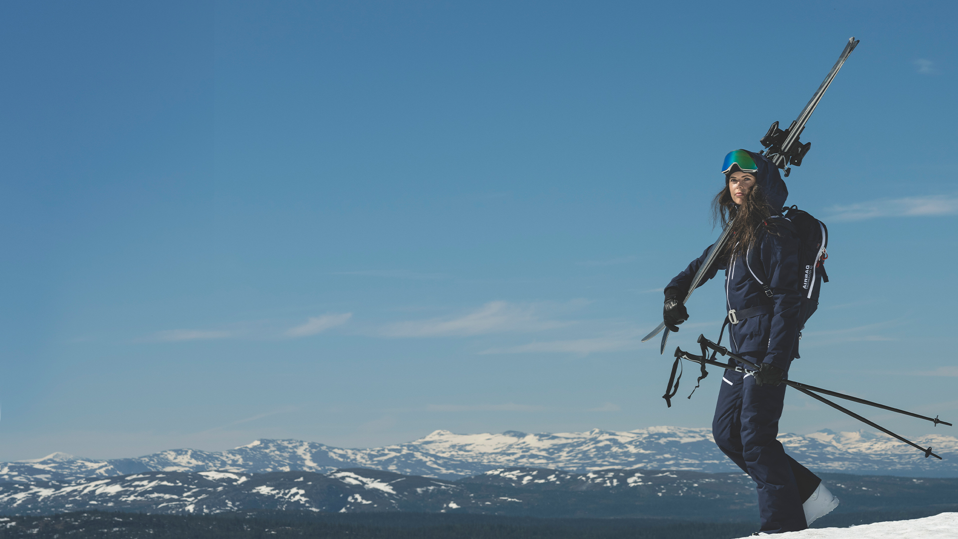 Kvinna i skidutrustning med skidor på axeln står på snötäckt fjälltopp med klar blå himmel i bakgrunden