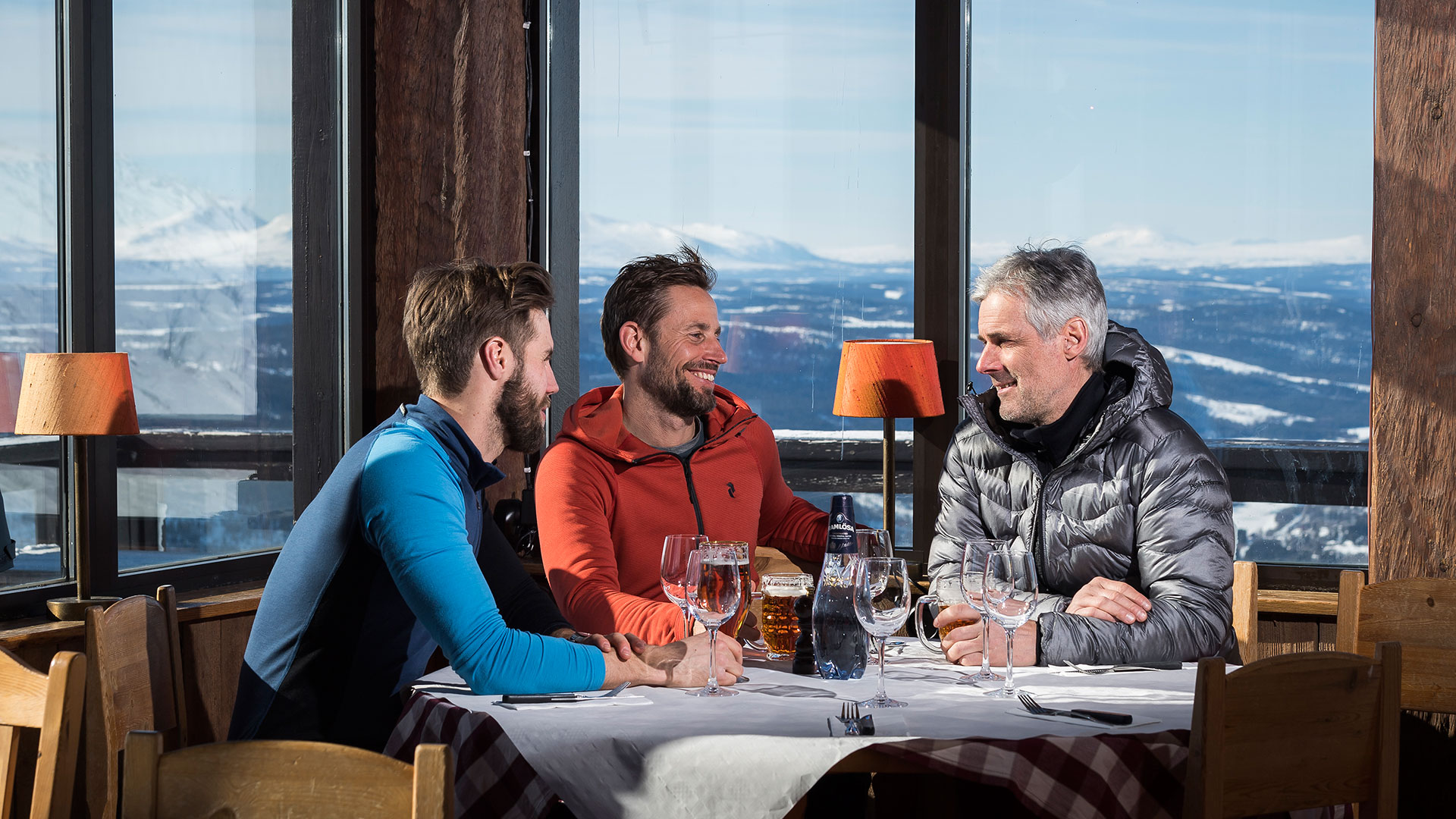 Tre män njuter av en måltid vid ett bord med panoramautsikt över snötäckta fjäll i en toppstuga