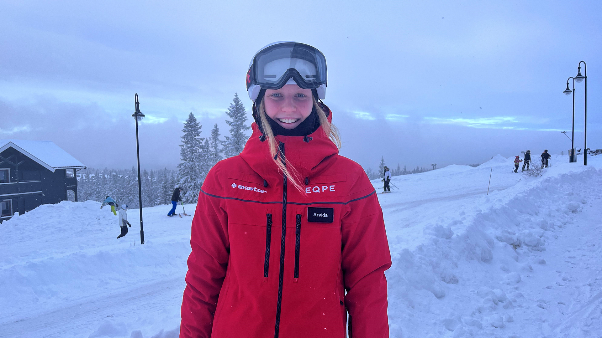 Person i röd skidjacka på vintrig skidort, omgiven av snö och skidåkare i bakgrunden