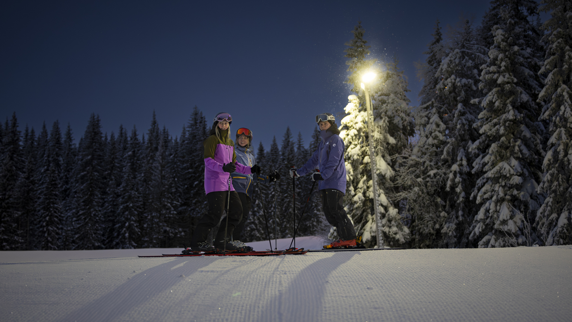 Tre skidåkare på en pist under kvällsåkning, omgivna av snötäckta träd och belysta av en lampa