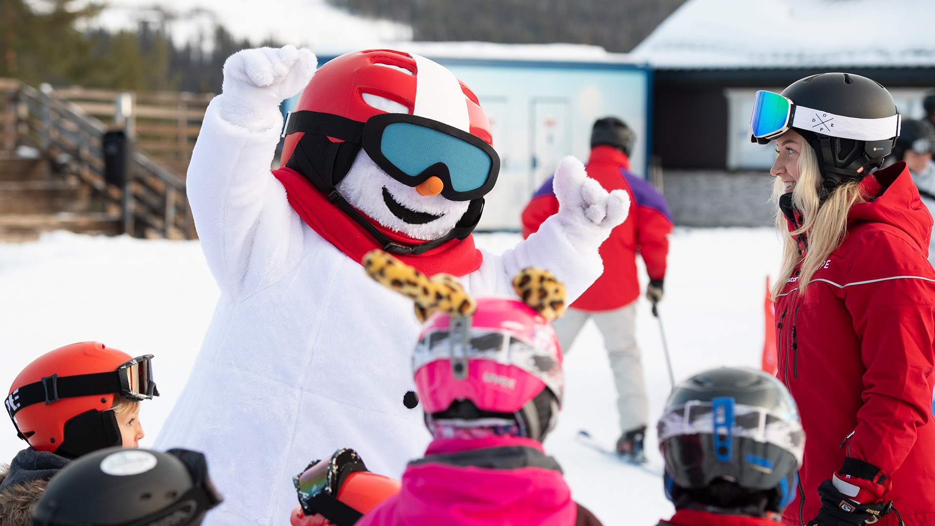 Snögubbe-maskot interagerar glatt med barn och skidlärare i röda vinterkläder på skidort i soligt väder