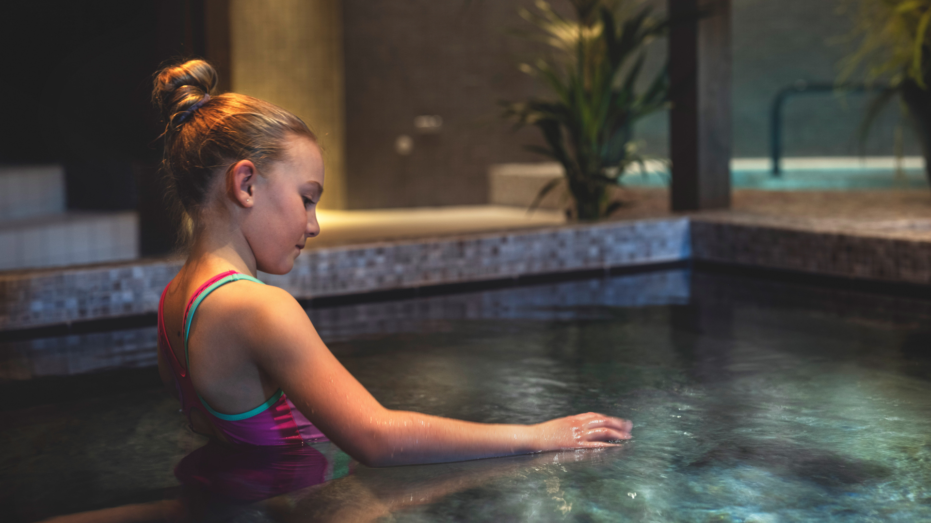 En ung flicka sitter vid en lyxig inomhuspool och rör vid vattnet, omgiven av en avkopplande spa-miljö
