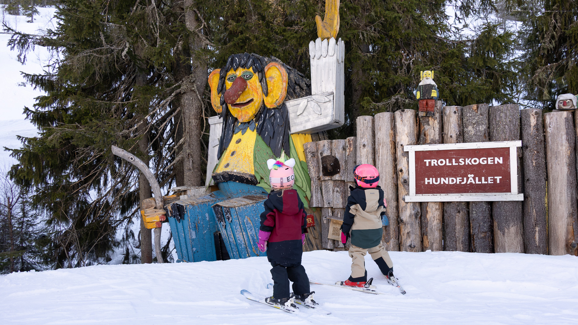 Barn på skidor vid trästaty av troll i Trollskogen, Hundfjället, omgiven av snö och skog