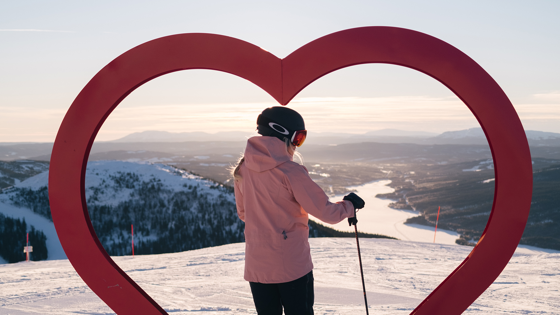 Person som åker skidor i fjällen med ett rött hjärta som inramning i förgrunden, snötäckta berg och dalar i bakgrunden under en klar himmel
