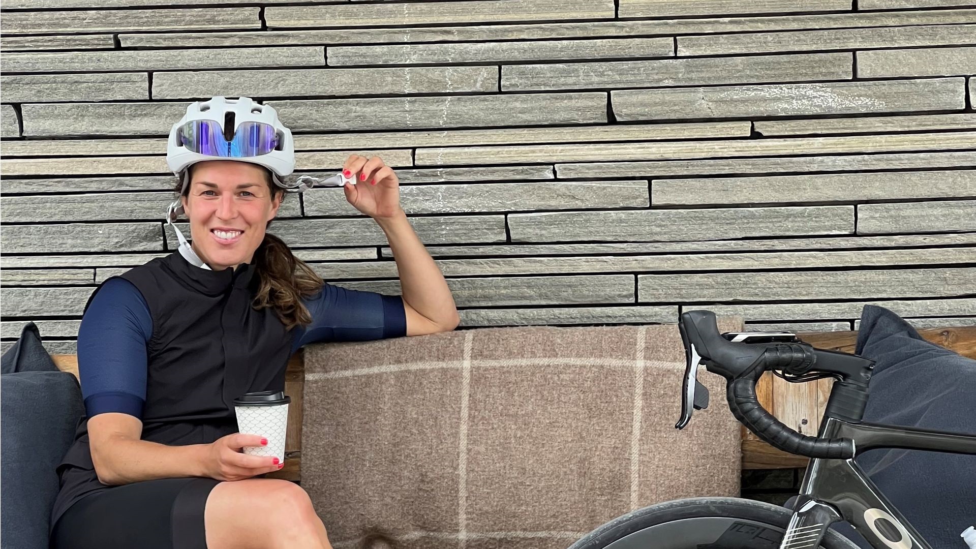 Cyklist med hjälm sitter på en soffa med en kaffekopp, cykel vid sidan om, framför en mur av gråa stenar