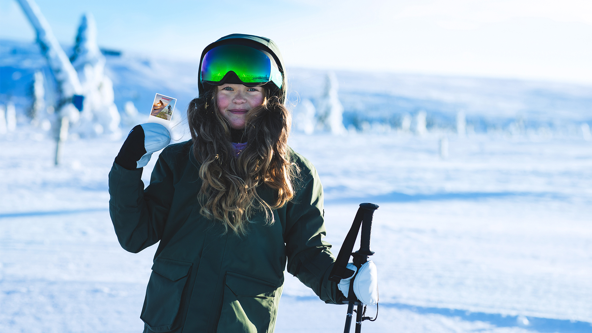 Young girl in a snow jacket and goggles holding a ski pole and a small photo, standing in a snowy landscape with frosted trees in the background
