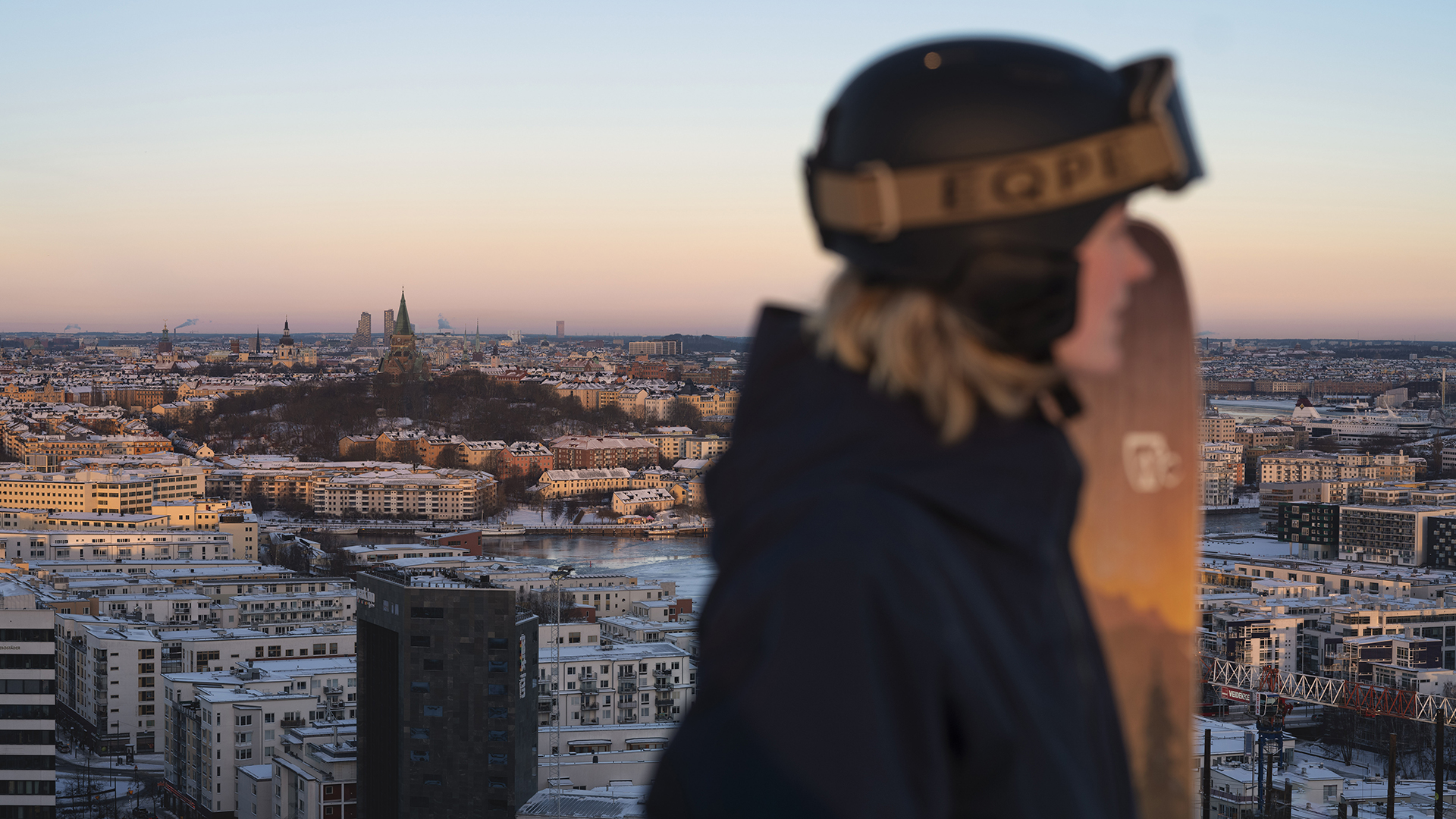 Person som bär hjälm med snowboard framför Stockholms stadslandskap vid solnedgång