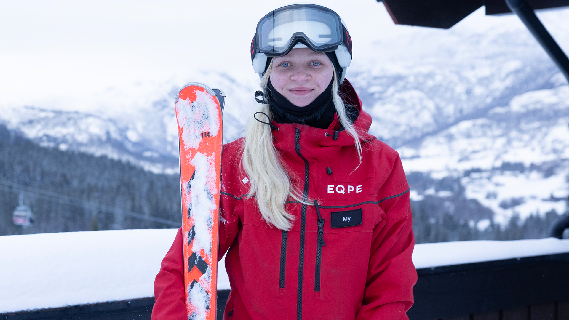Skidinstruktör i röd jacka med skidor stående framför snöklädda berg