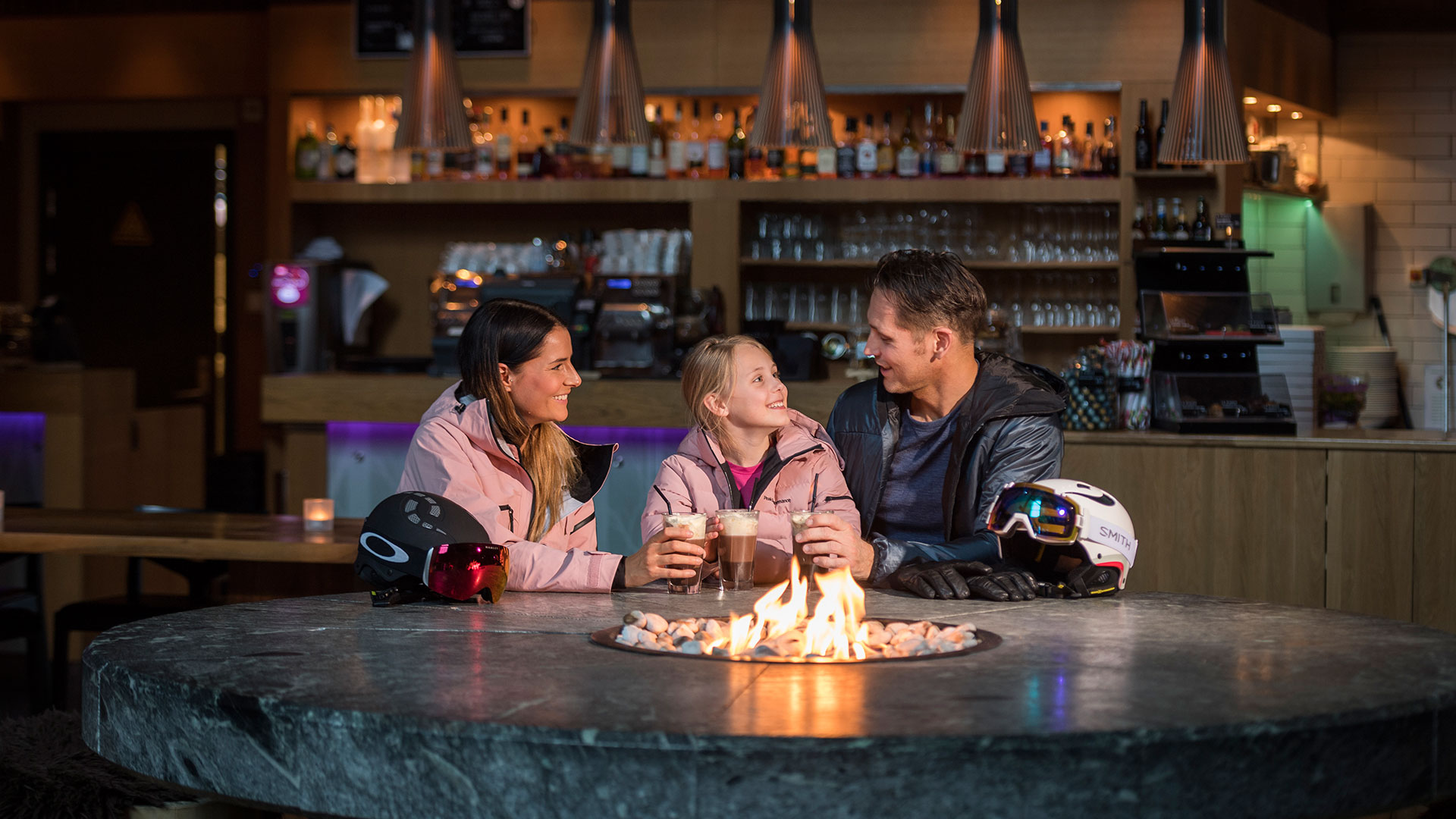 Familj sitter runt en eldstad inomhus på en skidort och dricker varma drycker, med skidhjälmar och glasögon på bordet. Restaurangmiljö i bakgrunden